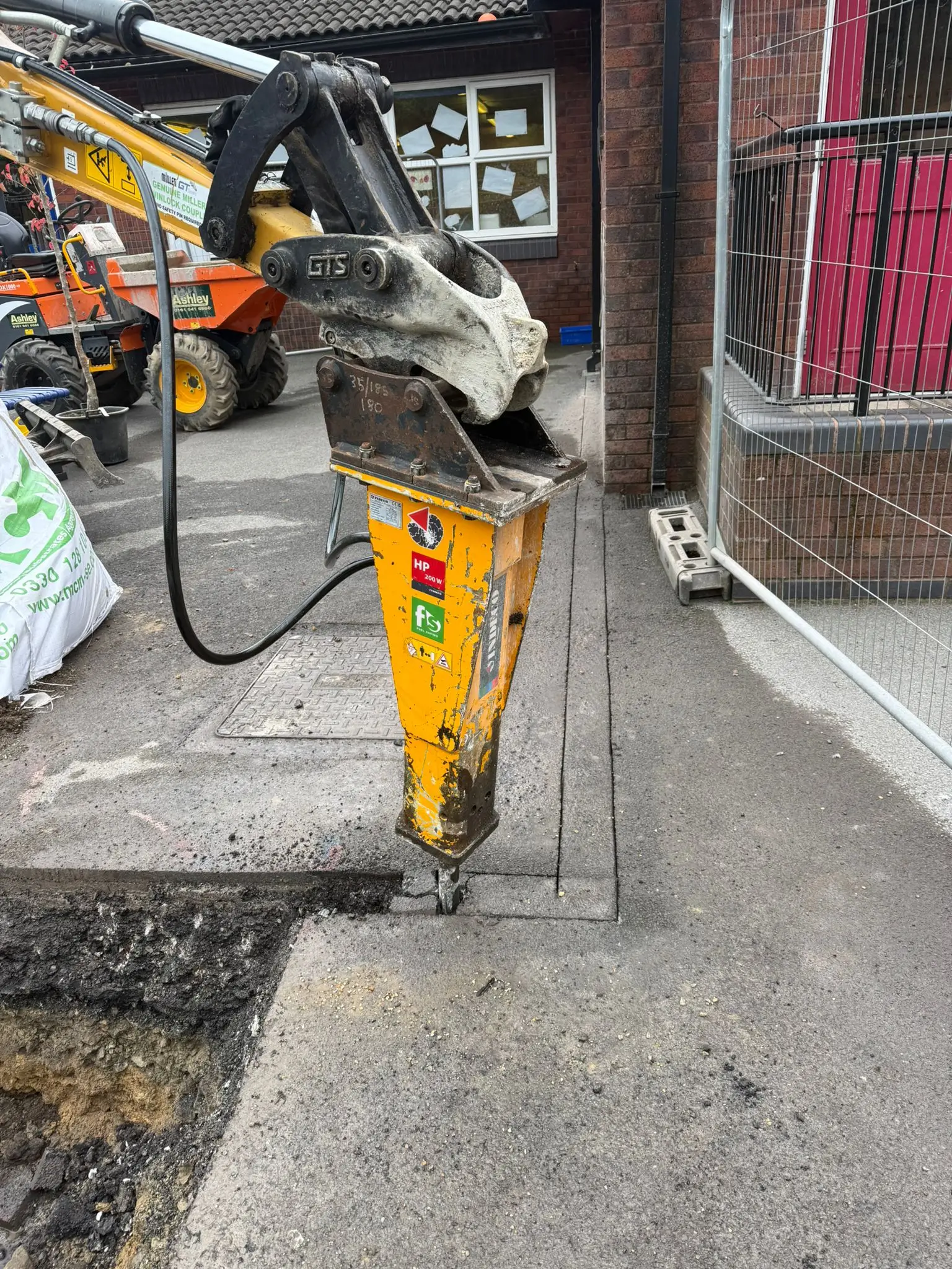 A yellow construction drill or core cutter stands on a sidewalk near a brick building, with construction equipment in the background.