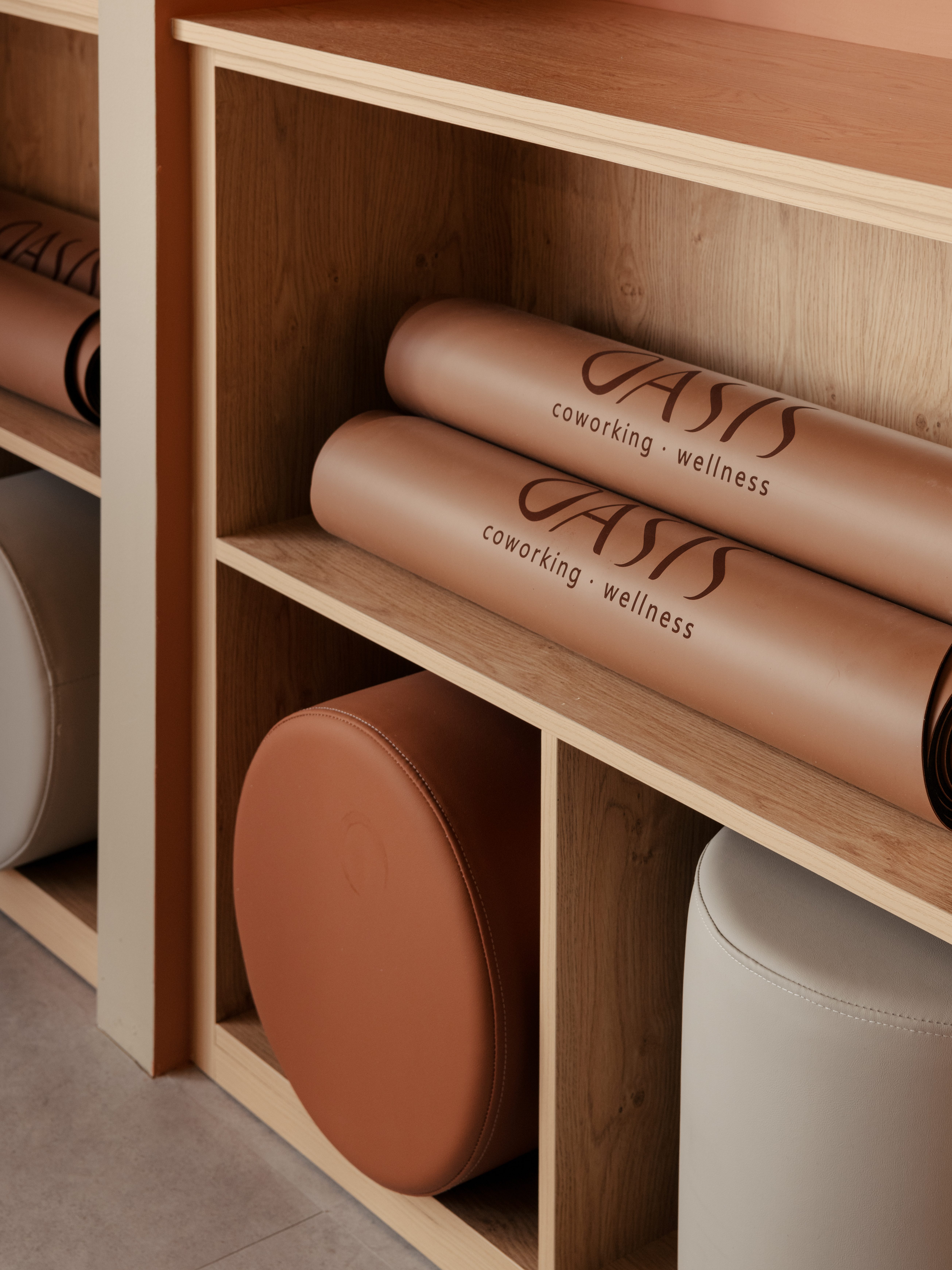 A shelf displaying rolled yoga mats labeled 'OASIS coworking - wellness' and cylindrical cushions, set against a neutral wall.