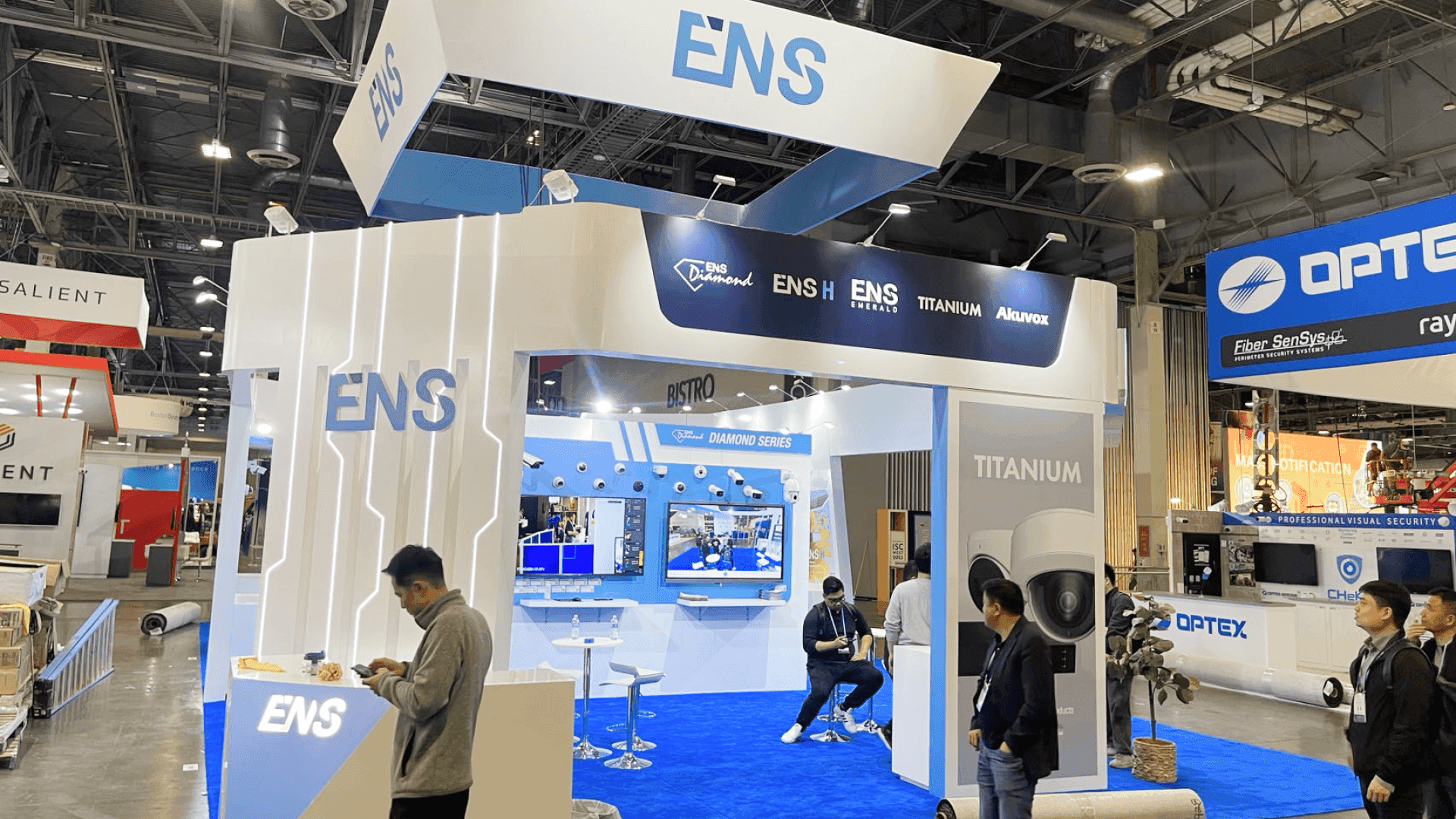 Side entry view of ENS ISC West 2025 booth with large overhead sign, illuminated product wall, blue flooring, and security technology display panels