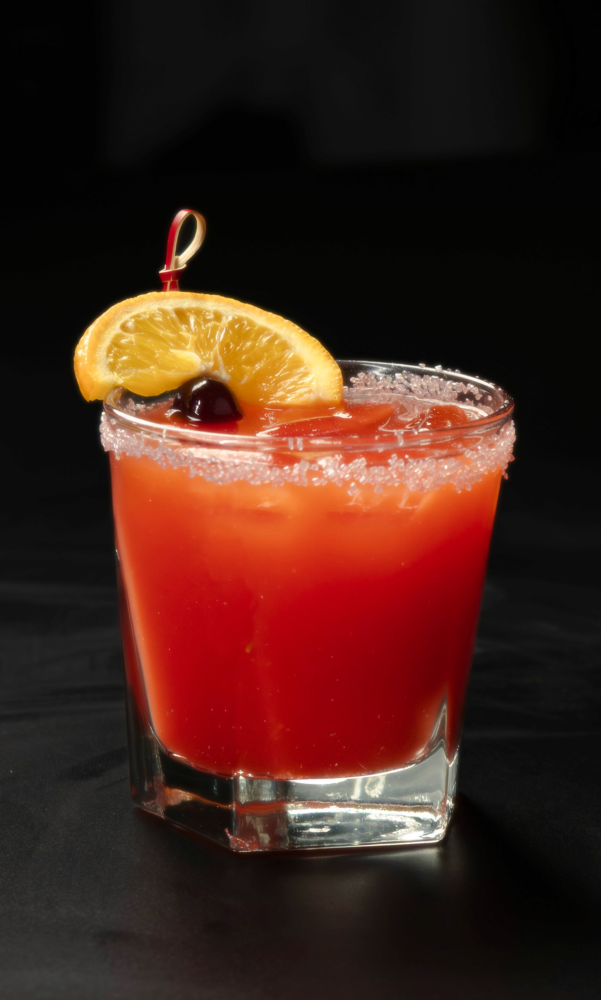 A vibrant orange tropical cocktail in a rocks glass rimmed with sugar, garnished with an orange slice and a maraschino cherry against a dark background.