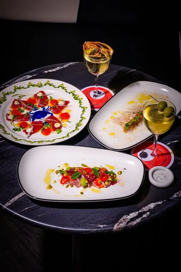 Plates of colorful crudo dishes topped with herbs, chili, and olive oil, served alongside a martini-style cocktail with green olives.