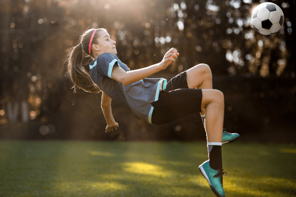 Image of a girl kicking a soccer ball and falling backwards onto the ground. She is wearing a soccer uniform and the sun is shining on her.