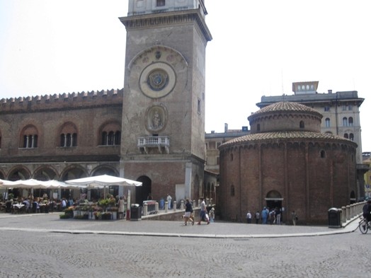 A photo of a paved square and some old stone buildings, one having a tower and a wall and the other circular.