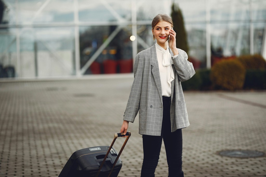 Stylish businesswoman speaking on smartphone while standing with luggage near airport