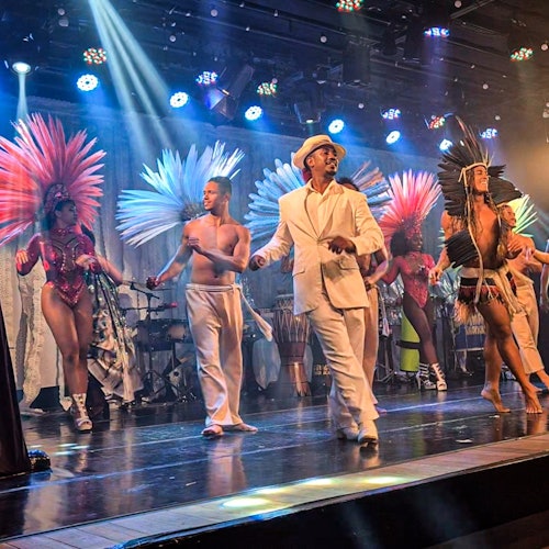 在充满活力的灯光下，身着色彩斑斓羽毛头饰的表演者和一名身穿白色西装和帽子的男子在舞台上翩翩起舞。