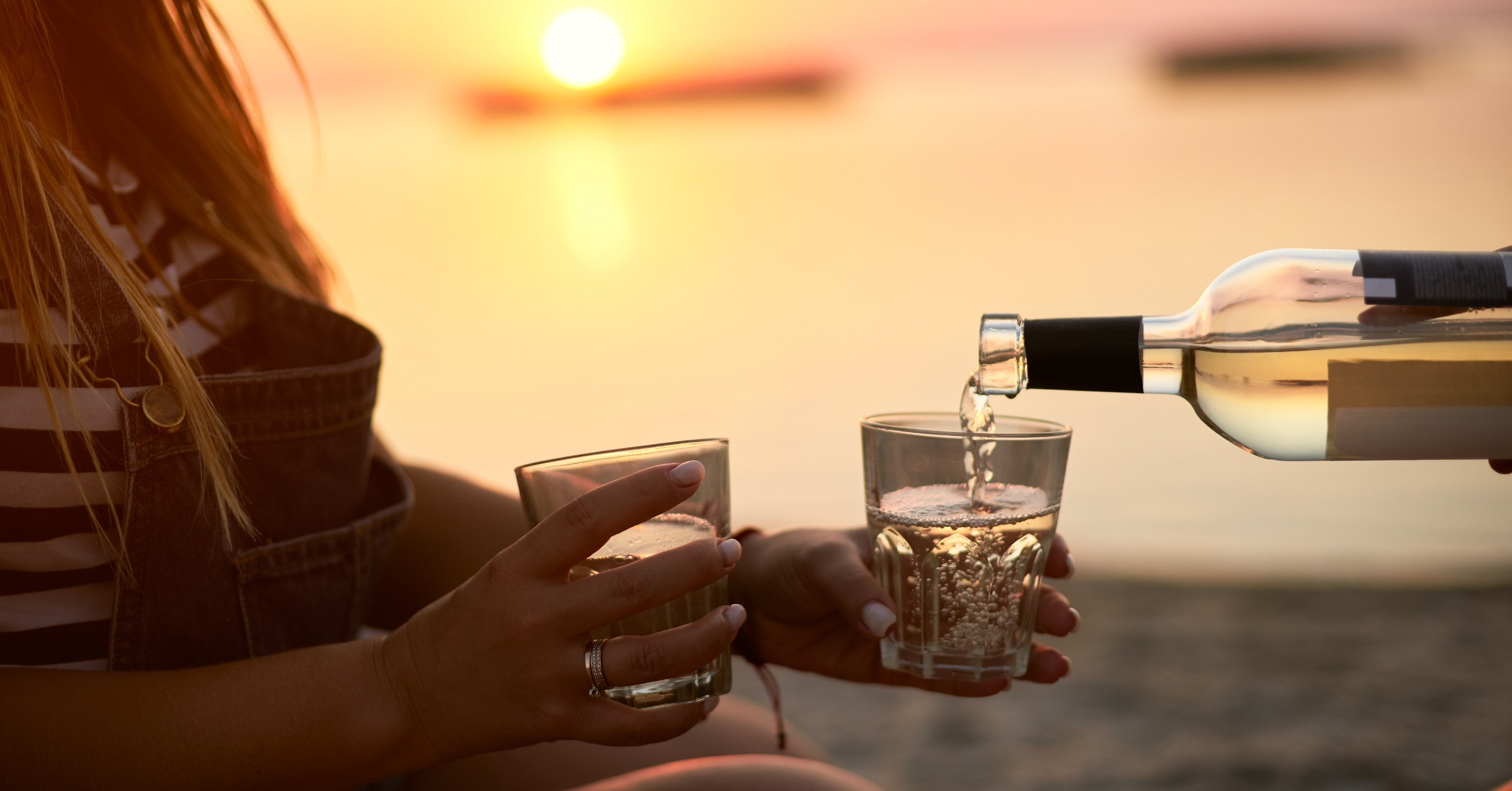 En kvinde står på en strand, med en flot solnedgang, holder to glas, mens der bliver hældt hvidvin op.