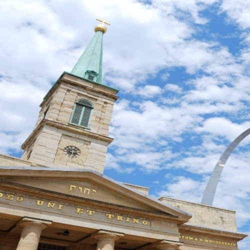 A historic church with a green steeple, clock, and cross under a partly cloudy sky, with part of a modern arch structure visible.