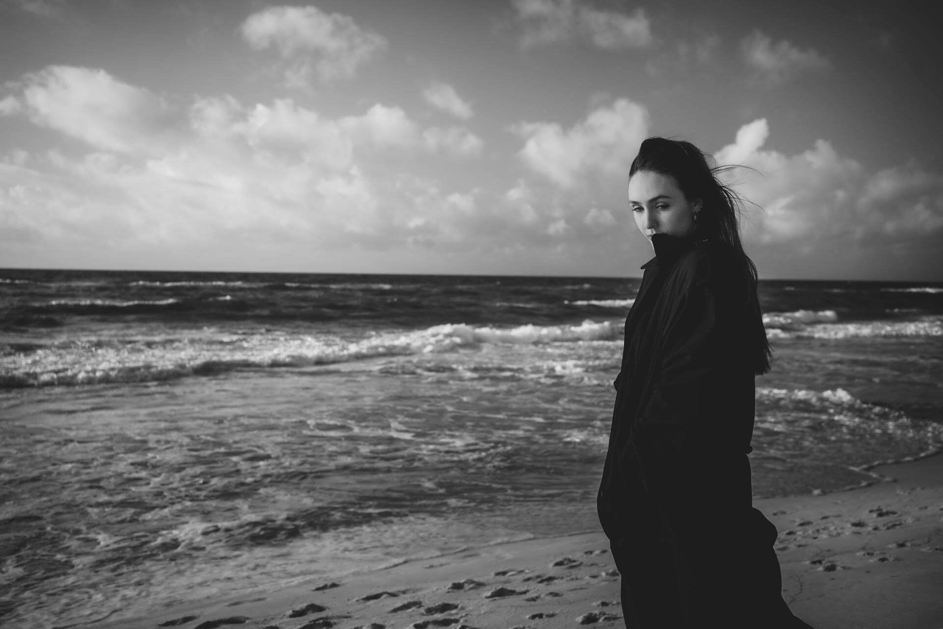 A woman in a black coat stands on the beach, gazing at the waves under a cloudy sky