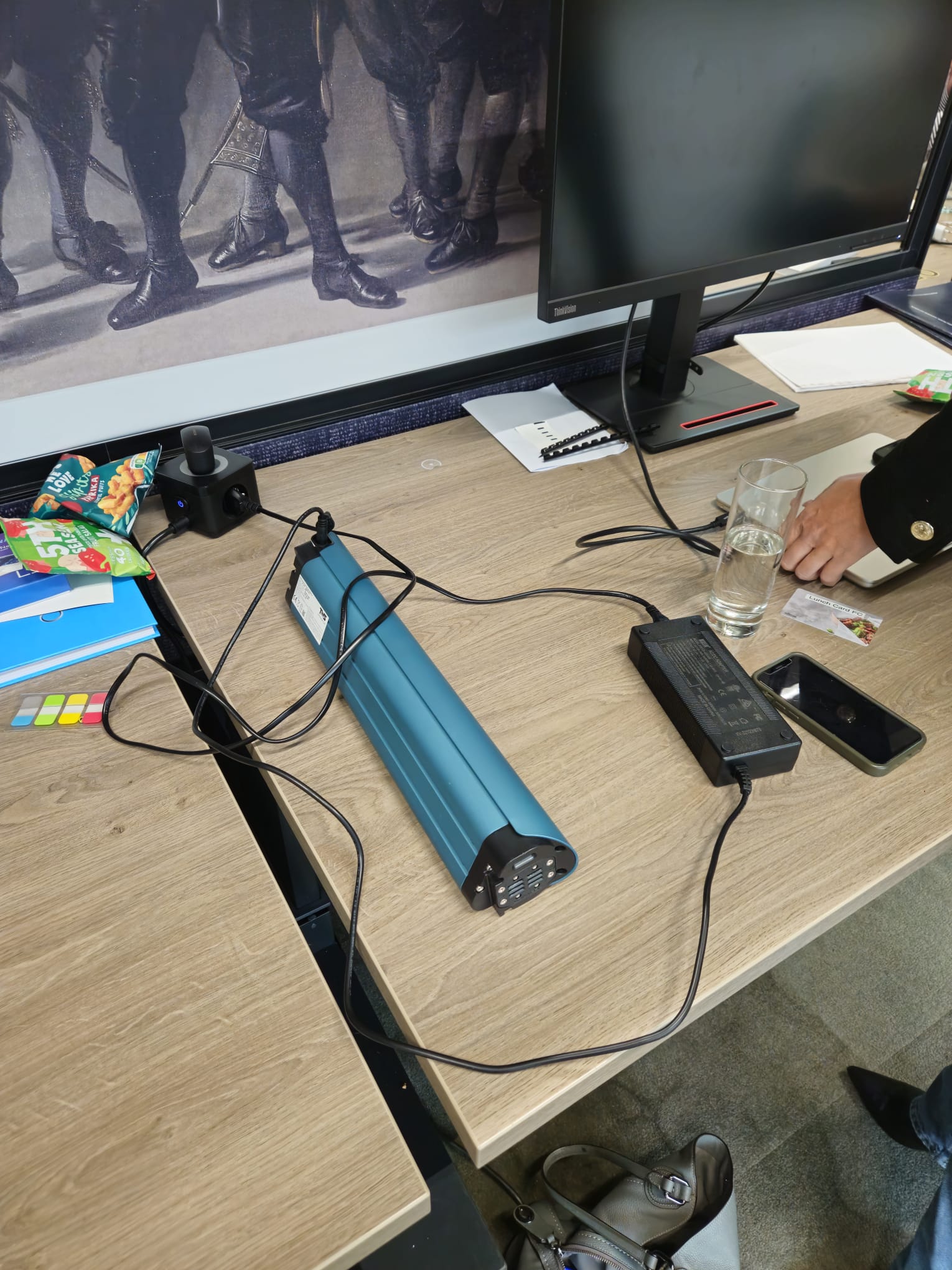 An e-bike battery charging on an office desk beside a colleague's workspace, with the charger and cable running across the desk and down to the floor.