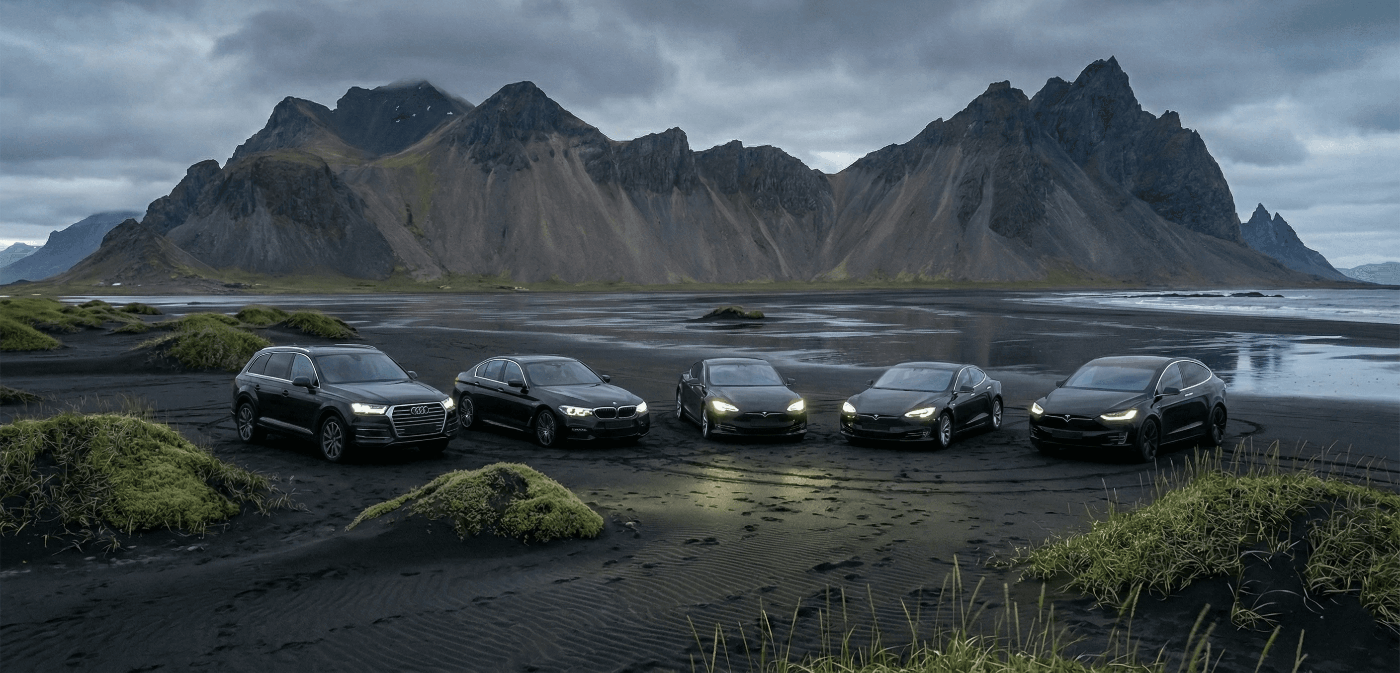 A fleet of five dark luxury cars, including Audis and Teslas, parked in a row on a black sand beach with jagged mountains in the background.