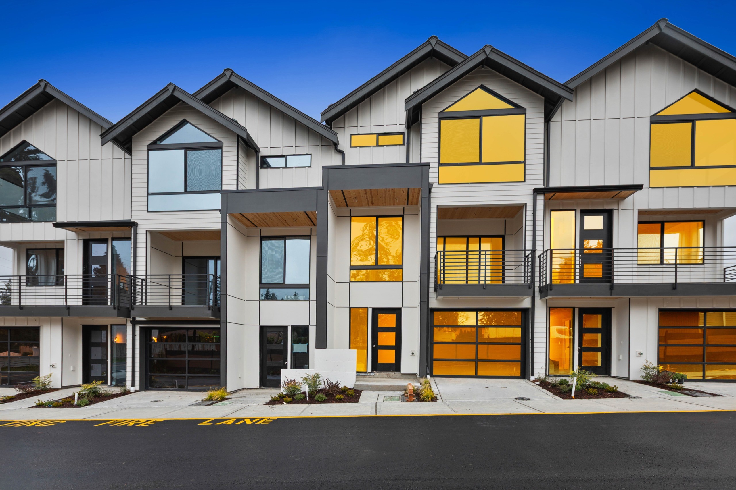 Street-facing twilight exterior of a modern luxury townhome row with wood-tone garage doors, grey board-and-batten siding, upper balconies, and glowing warm interior lighting