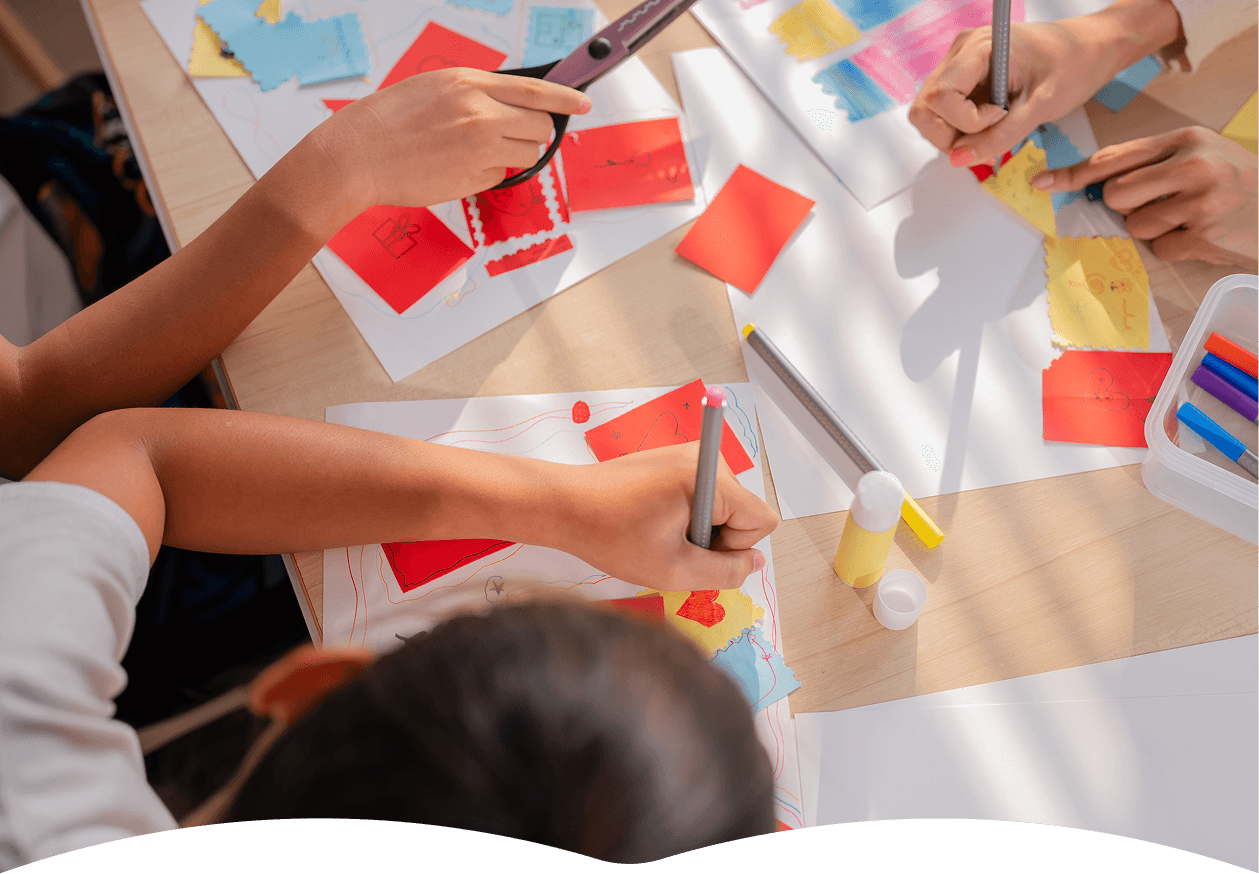Children are sitting at a wooden table, engaging in arts and crafts by cutting and coloring paper with crayons and scissors, surrounded by colorful paper pieces and glue sticks, illuminated by natural light.
