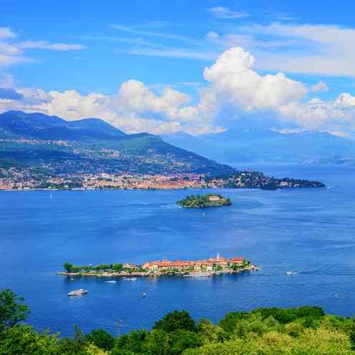 Aerial view of a lake with small islands, boats, and a coastal town, set against a backdrop of hills and a partly cloudy sky.