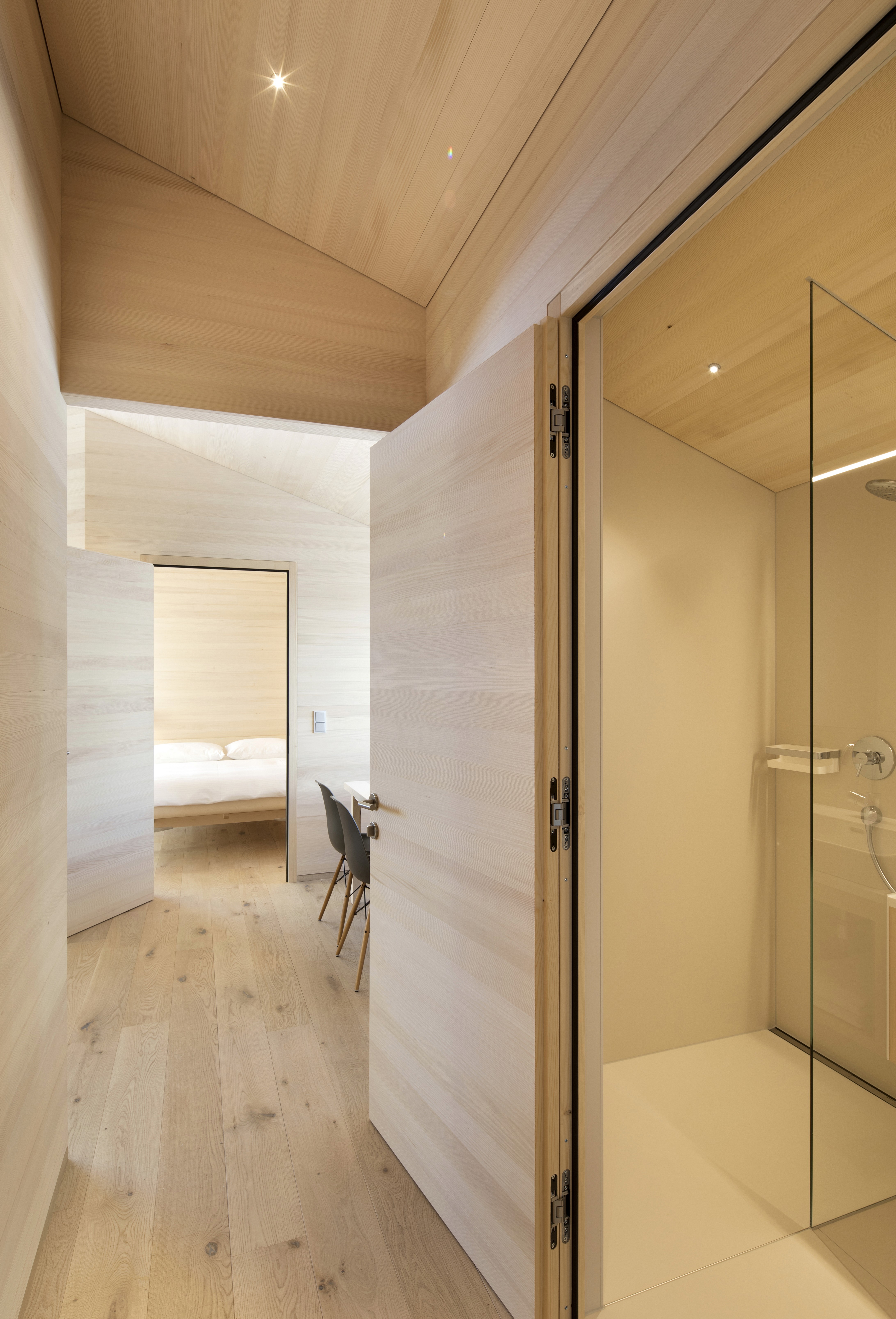 Light-wood hallway leading to a bedroom, with an open door revealing a modern glass-walled shower.