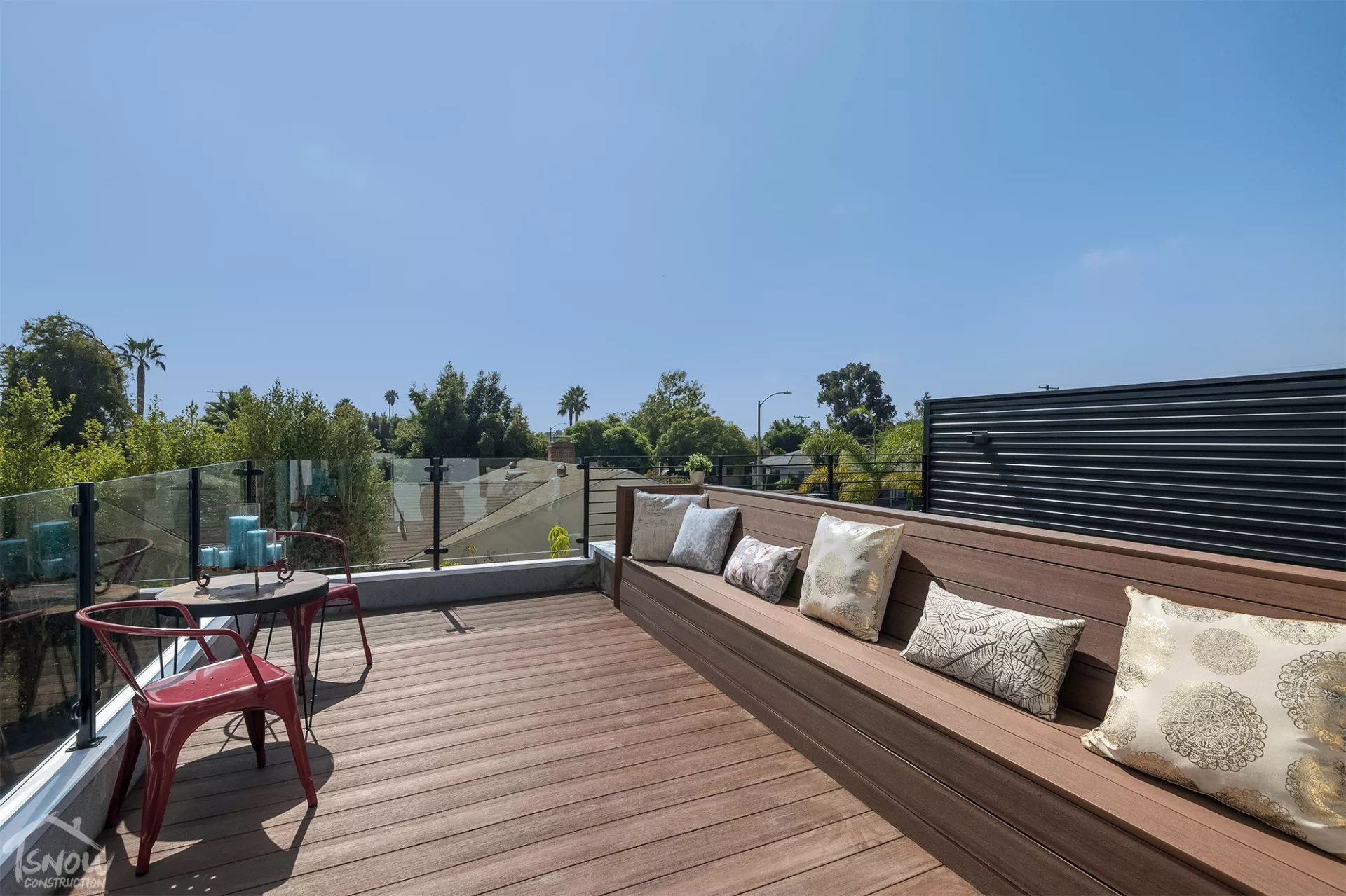 Rooftop deck build with wooden planks, cozy seating adorned with decorative pillows, and a view of lush greenery under a clear blue sky.