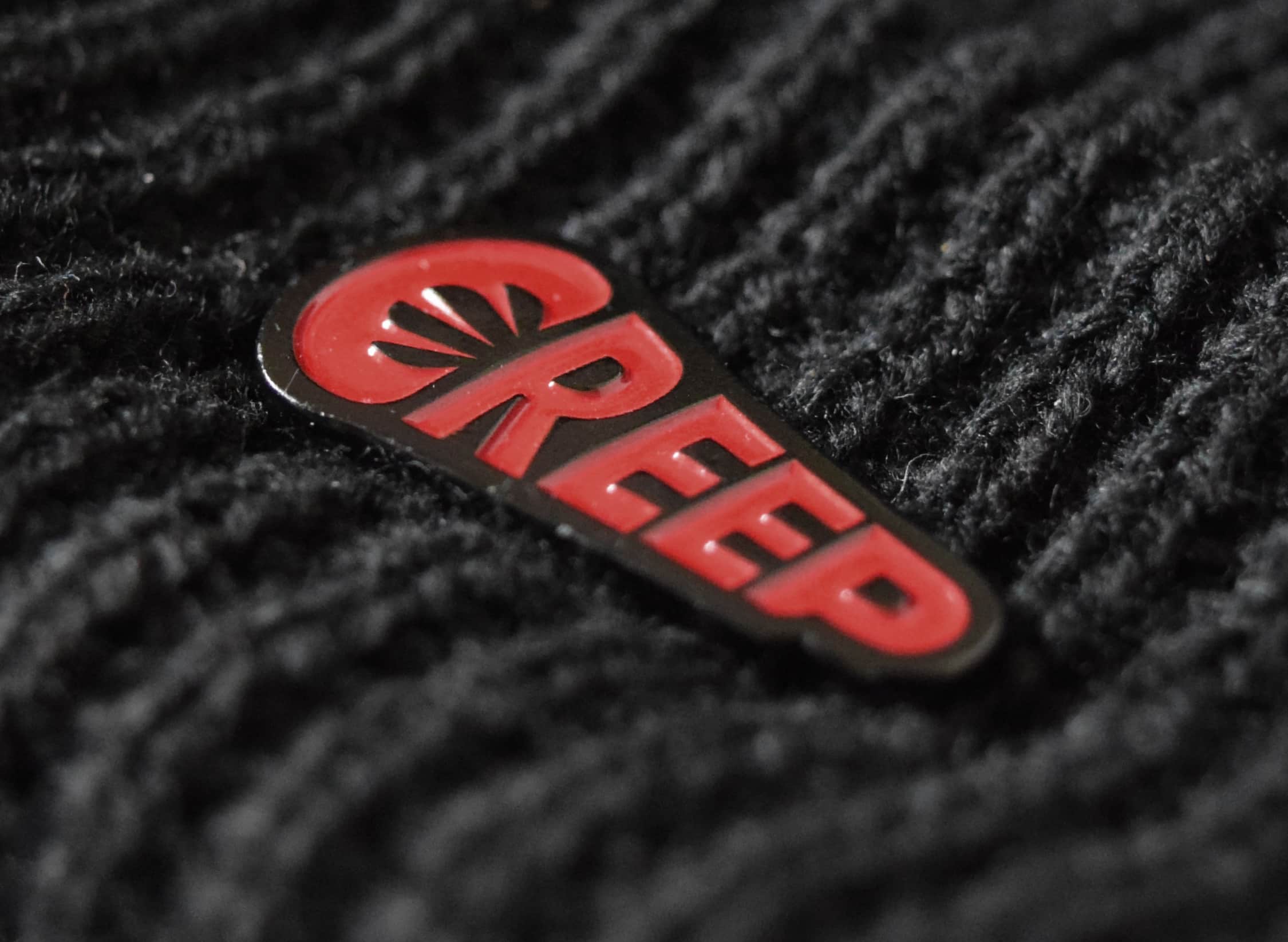 Red creep word-mark soft enamel pin badge on black beanie.
