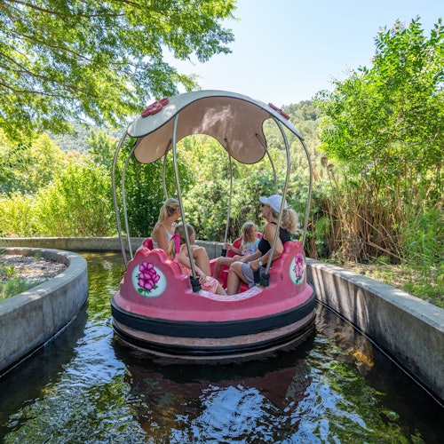 Empat orang menaiki bot pedal bertema bunga berwarna merah jambu di saluran air yang sempit dikelilingi oleh kehijauan yang subur pada hari yang cerah.