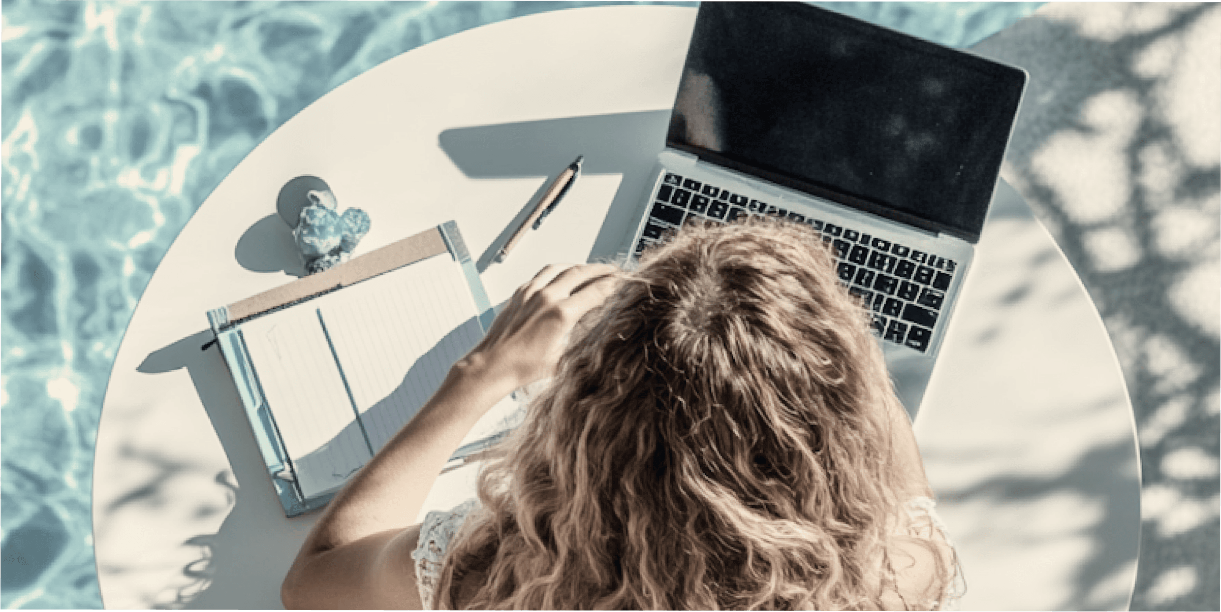 A person with curly blonde hair working on a laptop beside a pool, with a notebook and pen on the table.