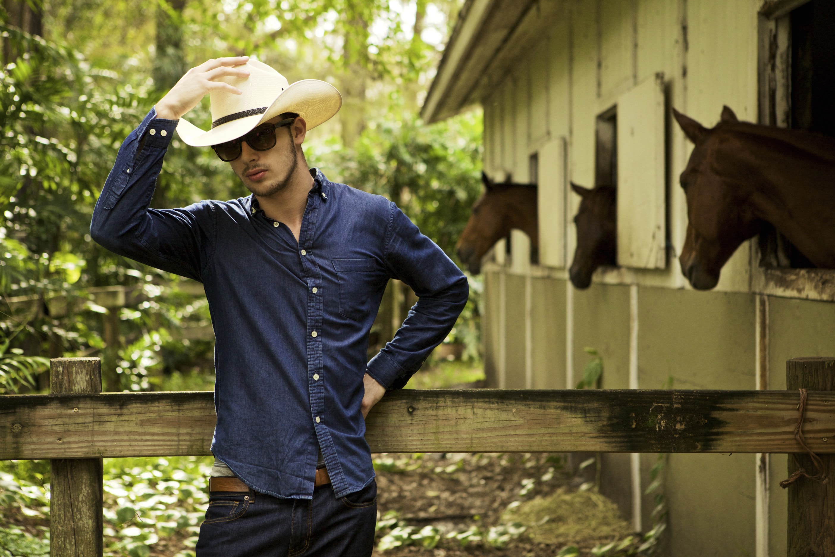 Model in a denim shirt and cowboy hat poses outdoors near a stable fence, Benjamin Jean lookbook photo.