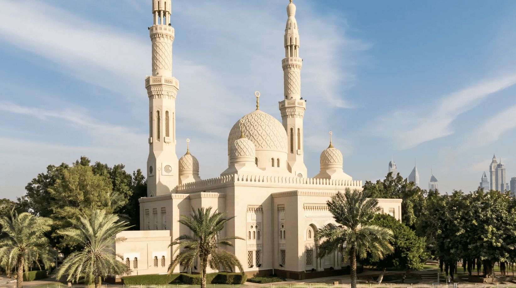 The large Jumeirah Mosque in Dubai surrounded by green grass and palm trees, set against a clear blue sky.