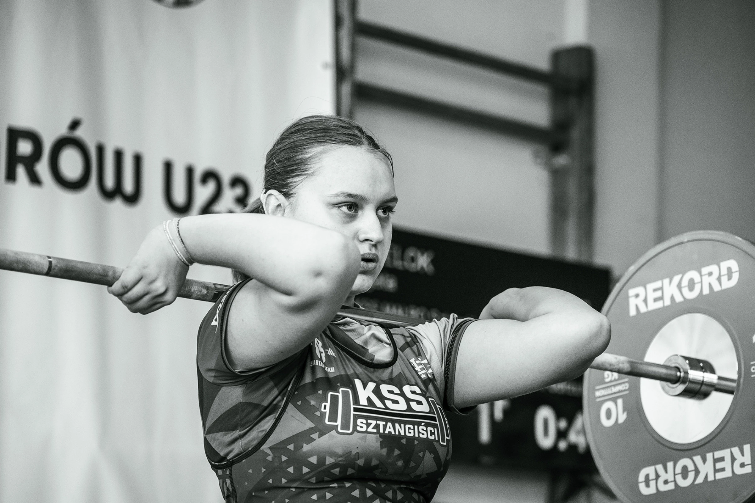 Młoda kobieta podnosi ciężar, trzymając sztangę przed ramionami. Zawody w podnoszeniu ciężarów U23.