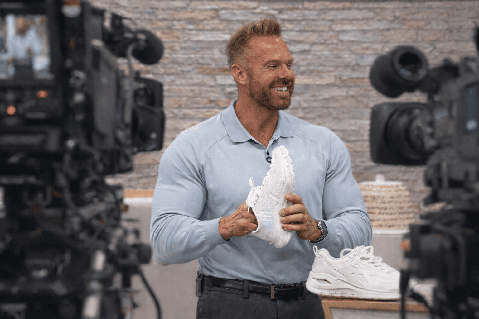 Mike Nicholls holding a white sneaker while presenting between television cameras.