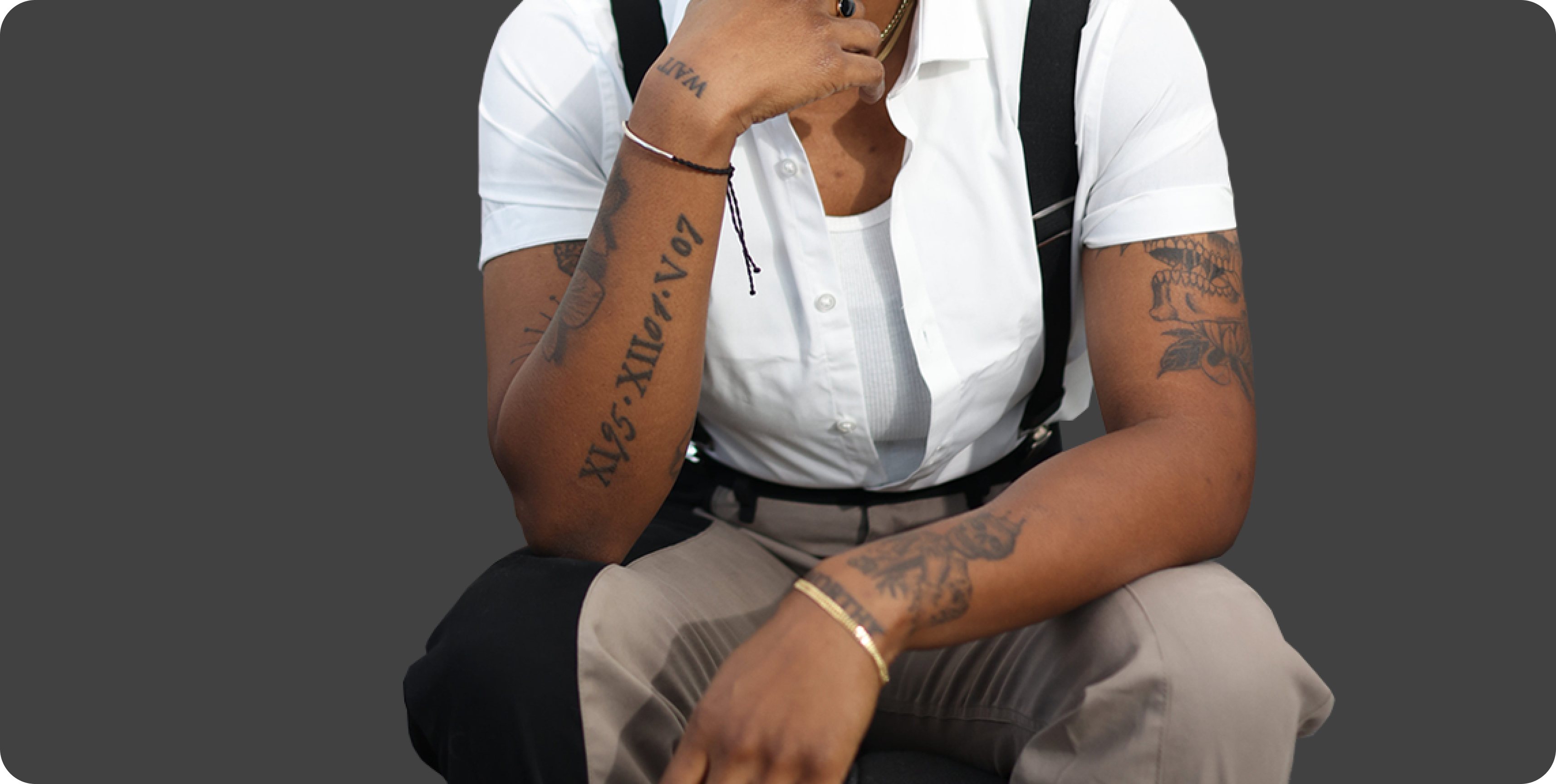 Ashleigh Warren, a  person in a white shirt and suspenders sits thoughtfully, showcasing arm tattoos against a black background.