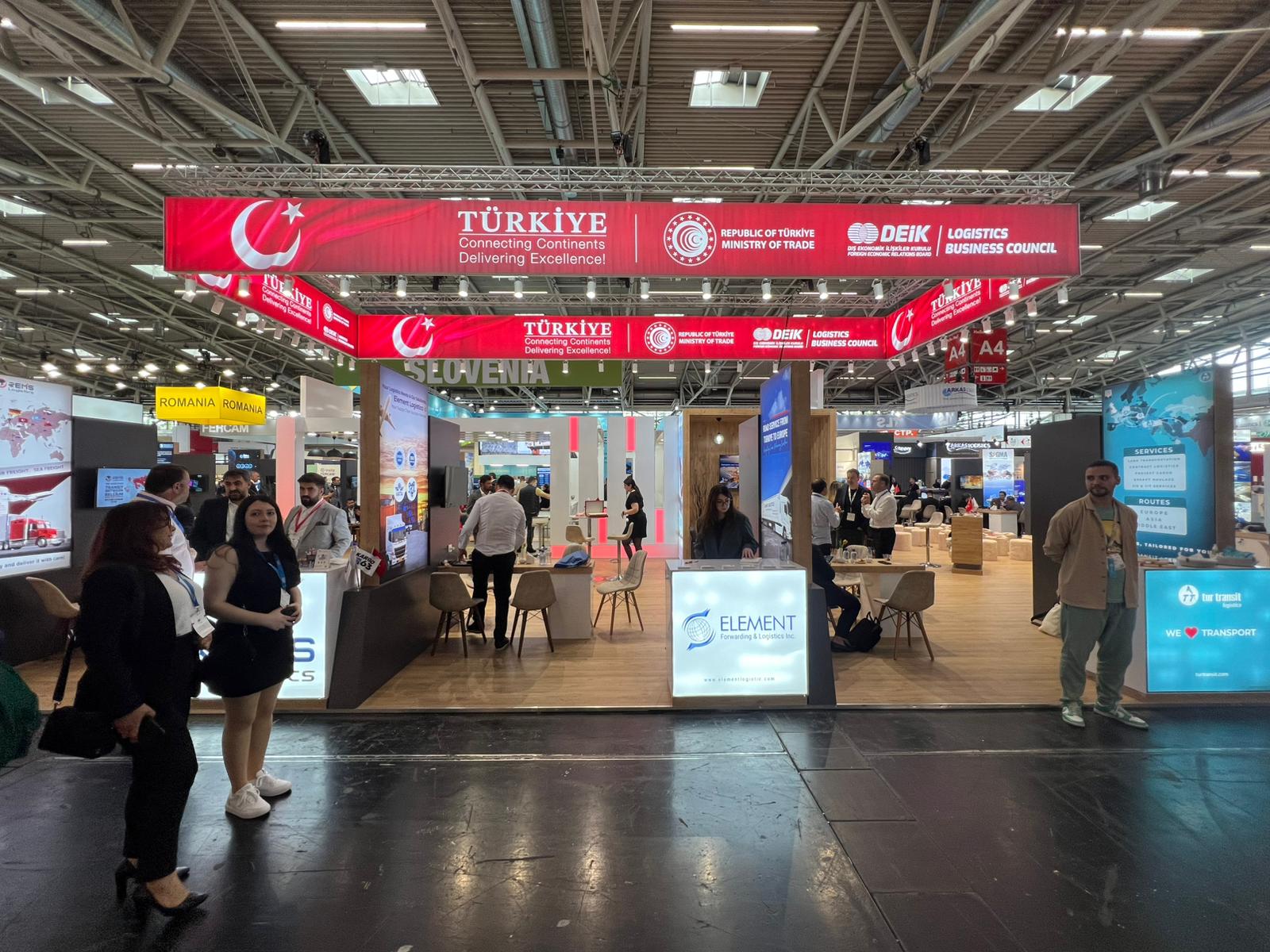 Alternate view of the DEİK pavilion layout, showing the wooden flooring, dark partition walls, and illuminated counters.