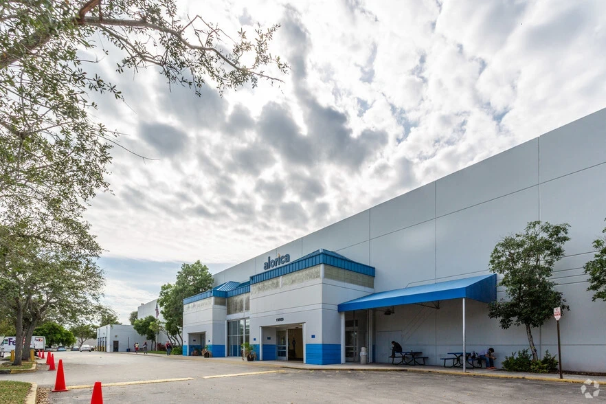 Spacious commercial building with blue accents under a cloudy sky. Trees and red cones line the sidewalk. 