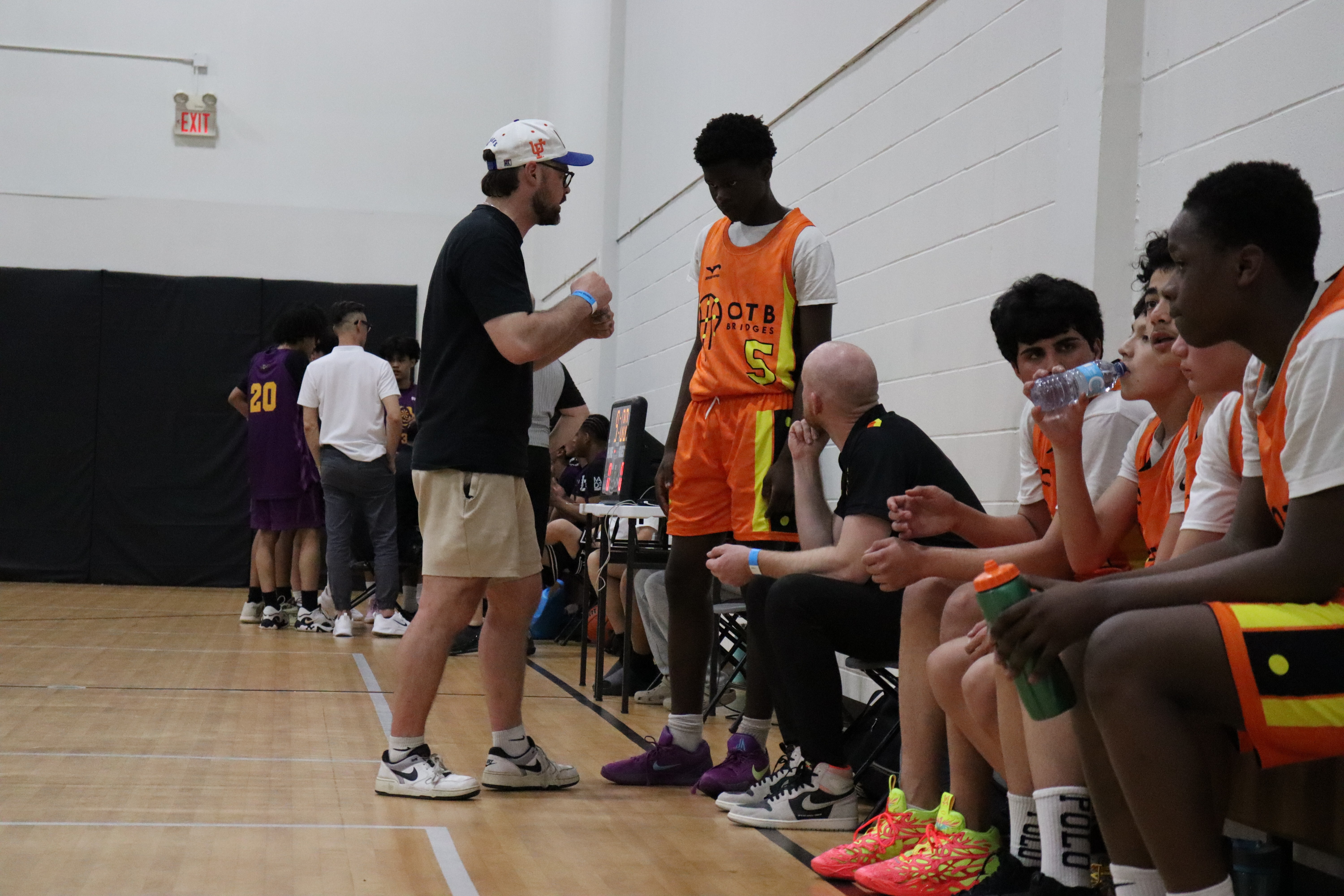 A youth basketball player in an orange jersey dribbling aggressively past a defender during a game, with a referee and spectators visible in the background on an indoor court.