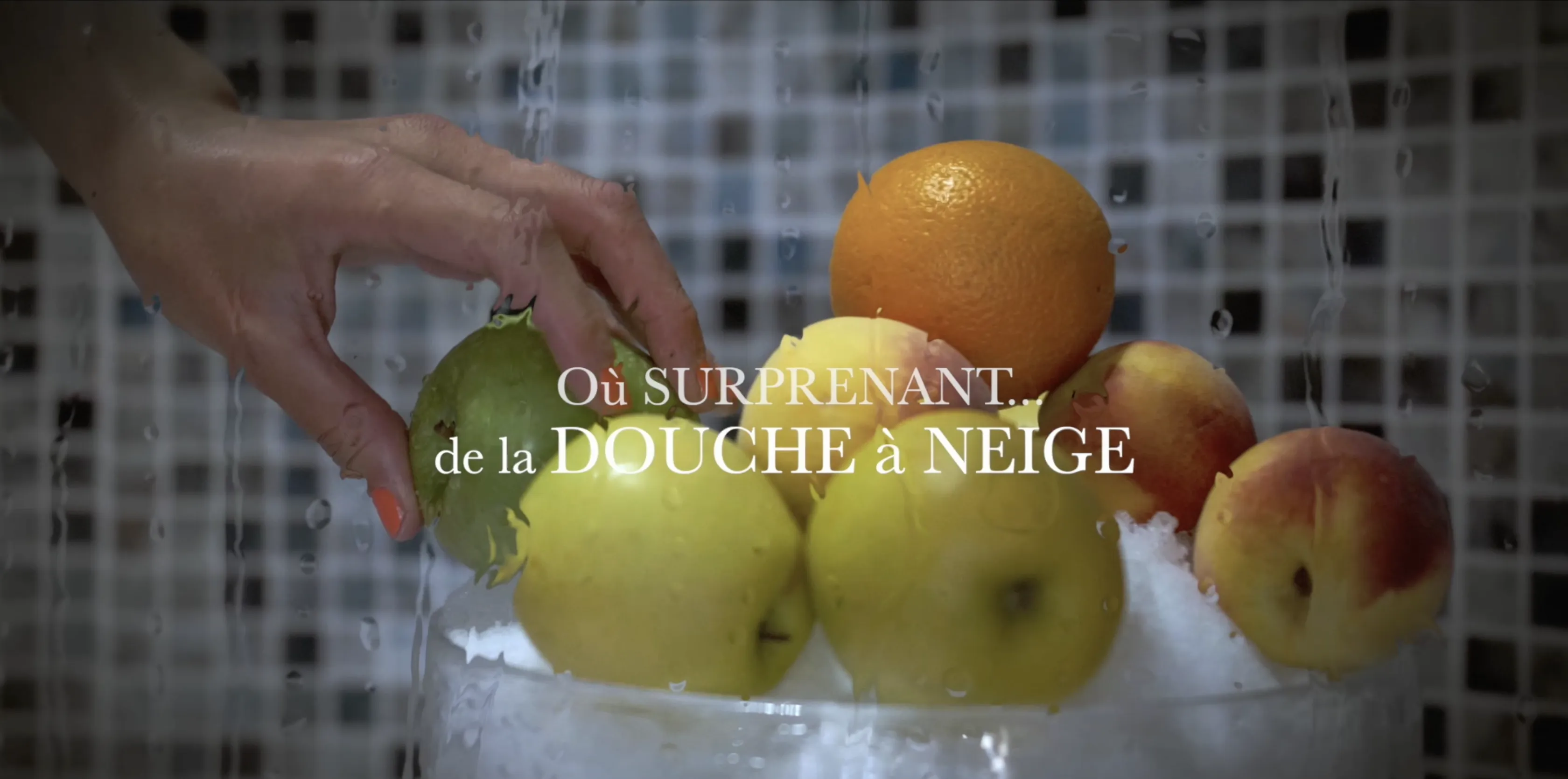 A hand reaches for a lime nestled among oranges, apples, and peaches in a glass bowl filled with ice, set against a tiled backdrop.