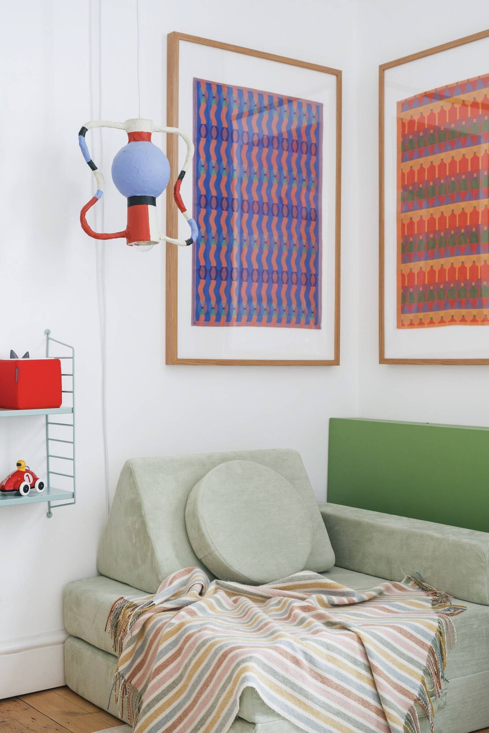 A living room corner with a vintage green sofa, a striped blanket, colorful contrasting abstract artwork on the walls, and a unique hanging lamp.