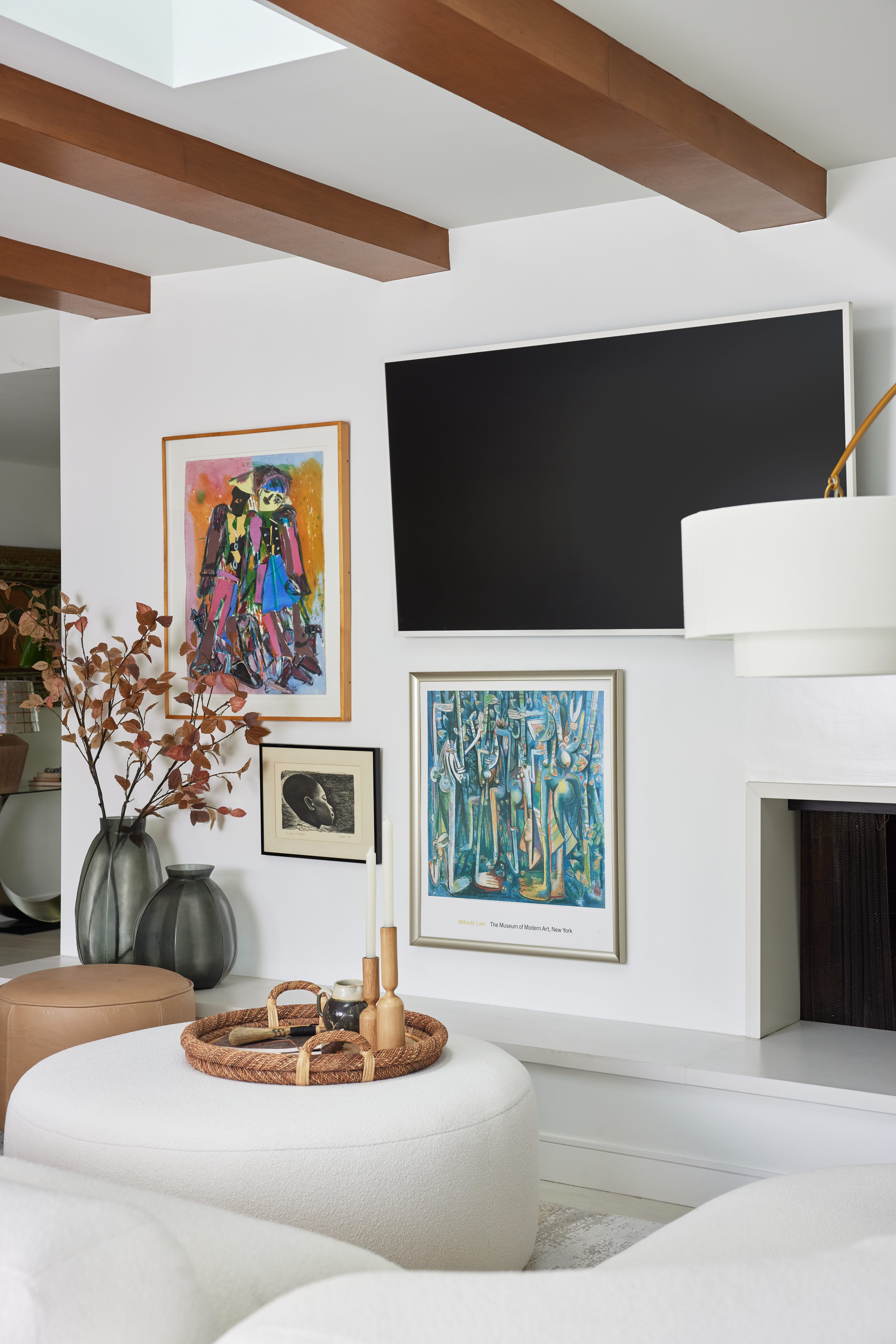 Bright family room with exposed beams, off-set fireplace, white boucle modular sofa, round tan ottomans, and heritage art pieces by Jubilee Interiors in Los Angeles, California