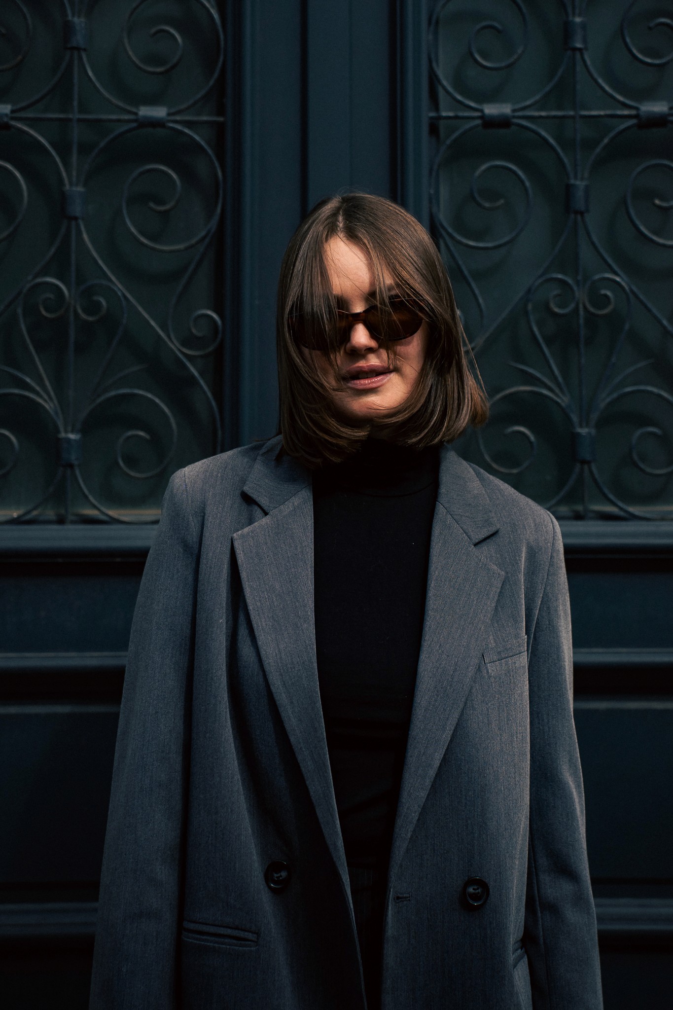 Portrait d’une femme debout à une porte d’entrée noire lors d’un shooting photo en extérieur, regardant dans la rue, photographie de portrait par Robin Masson