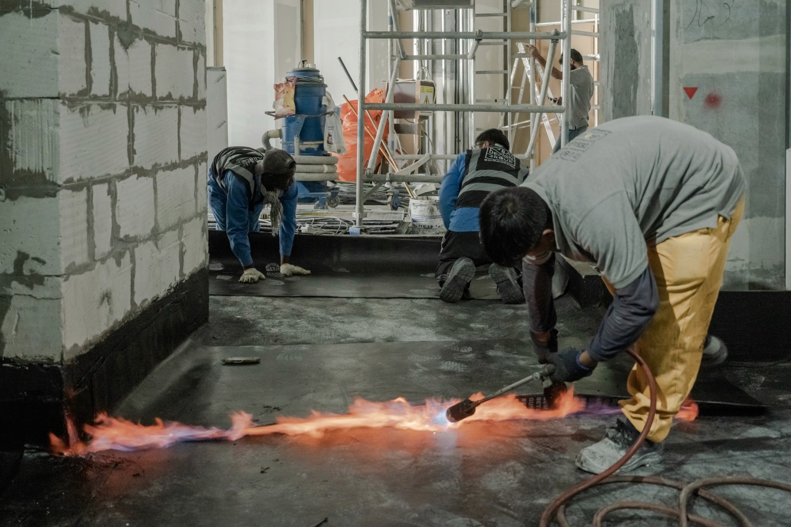 Workers in a construction site are engaged in waterproofing the floor with a torch, surrounded by scaffolding, building materials, and unfinished walls, demonstrating industrial labor in progress.