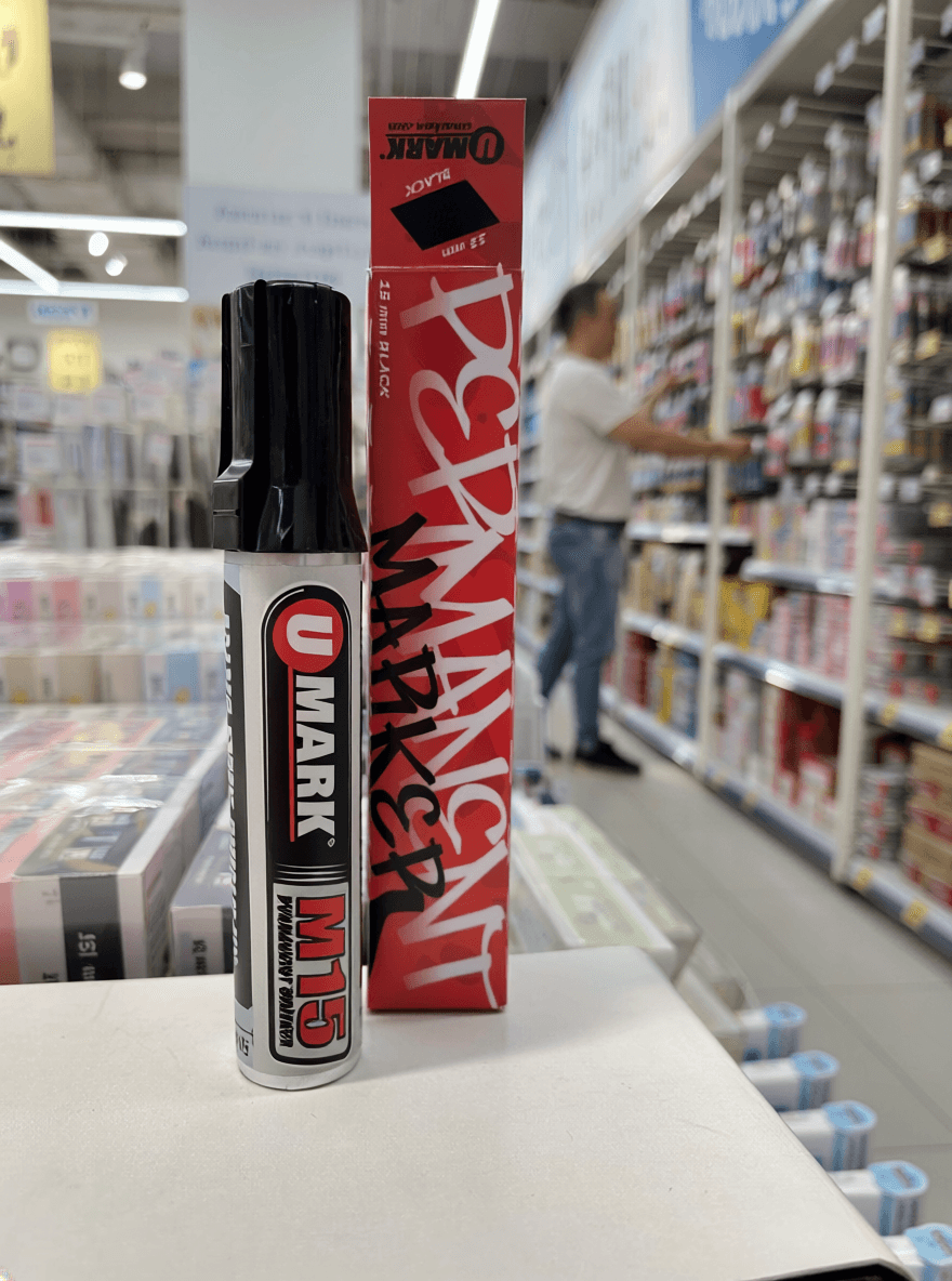 A black permanent marker and its red box labeled “U Mark” stand upright on a display table in an art supply store. The box features bold white graffiti-style lettering and the word “black” near the top. In the softly blurred background, a person browses shelves filled with colorful art supplies under bright store lighting.