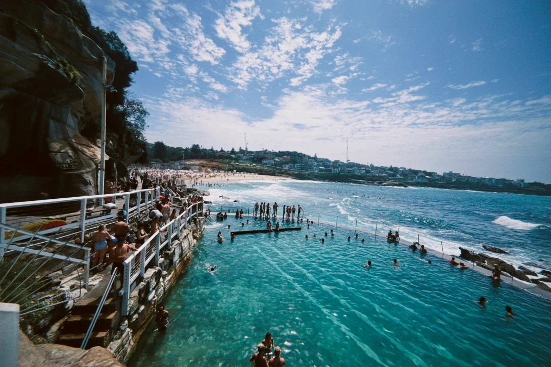 Scenic view of a crowded beachfront ocean pool with turquoise water, surrounded by rocky cliffs and a bustling beach, set against a backdrop of a clear blue sky and distant cityscape.