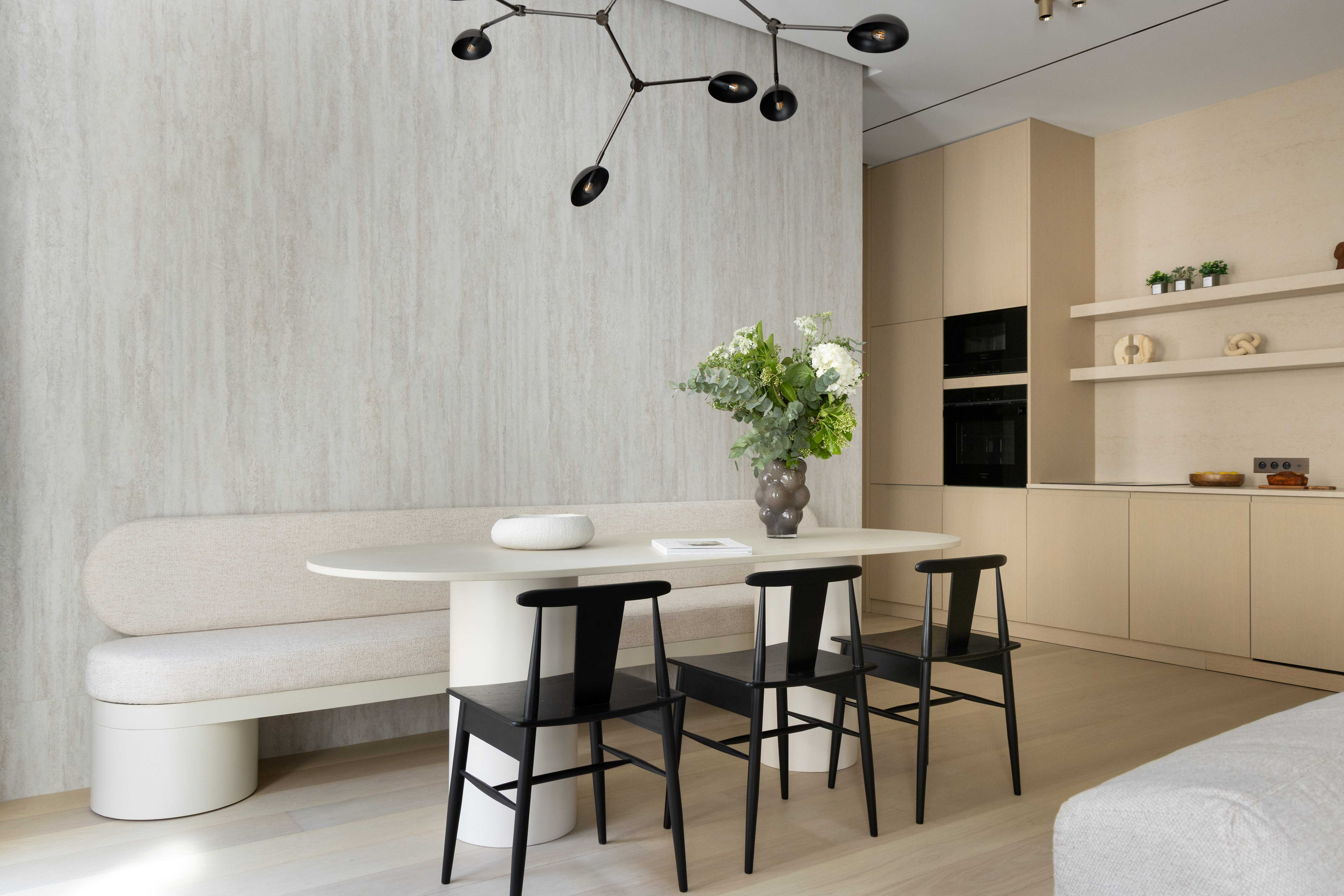 Coin repas avec table ovale beige, banquette courbe, chaises noires, suspension contemporaine et mur texture gris