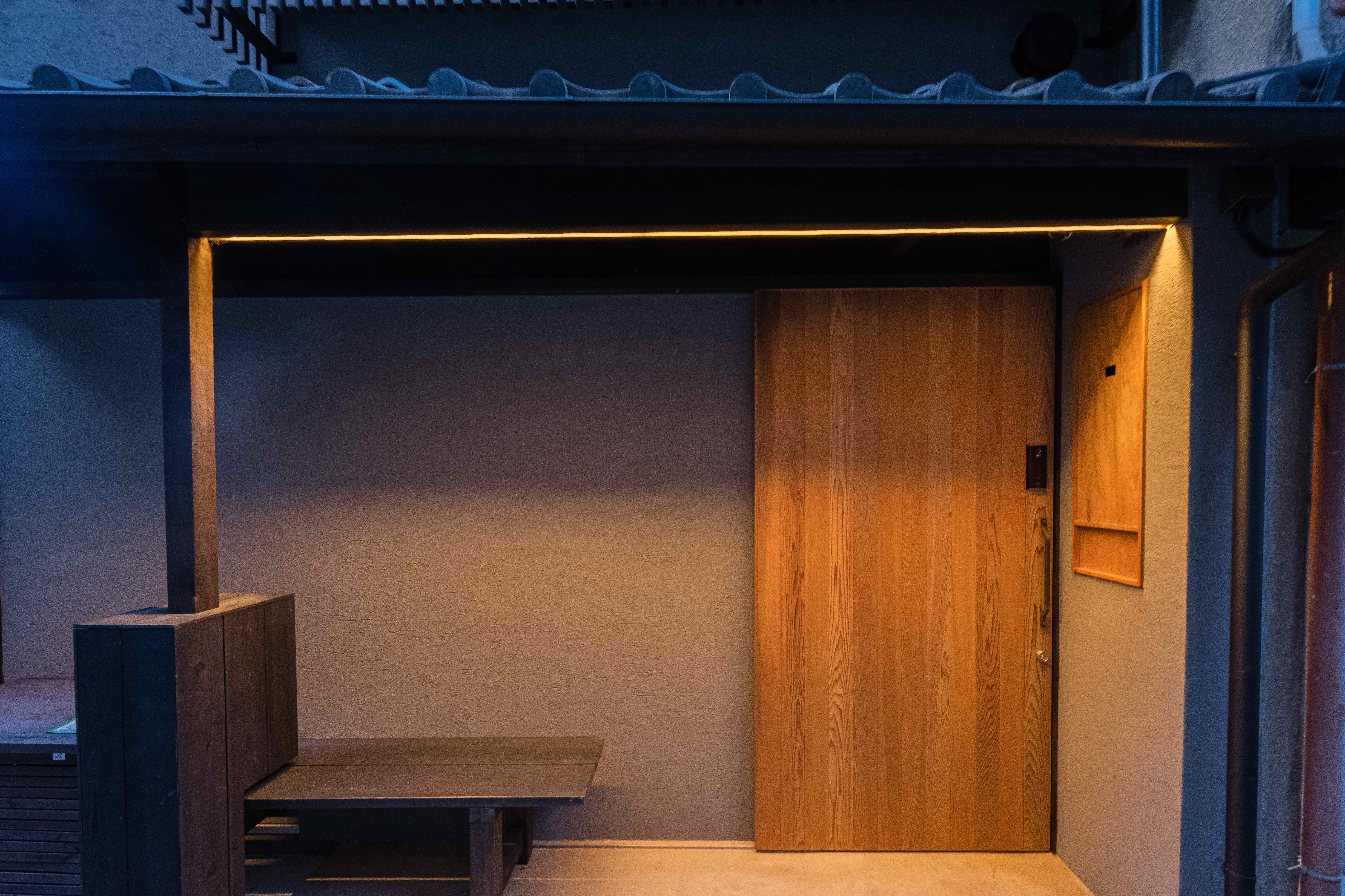 A cozy, modern entrance featuring a wooden door under a minimalistic awning with soft, warm lighting, creating an inviting atmosphere.