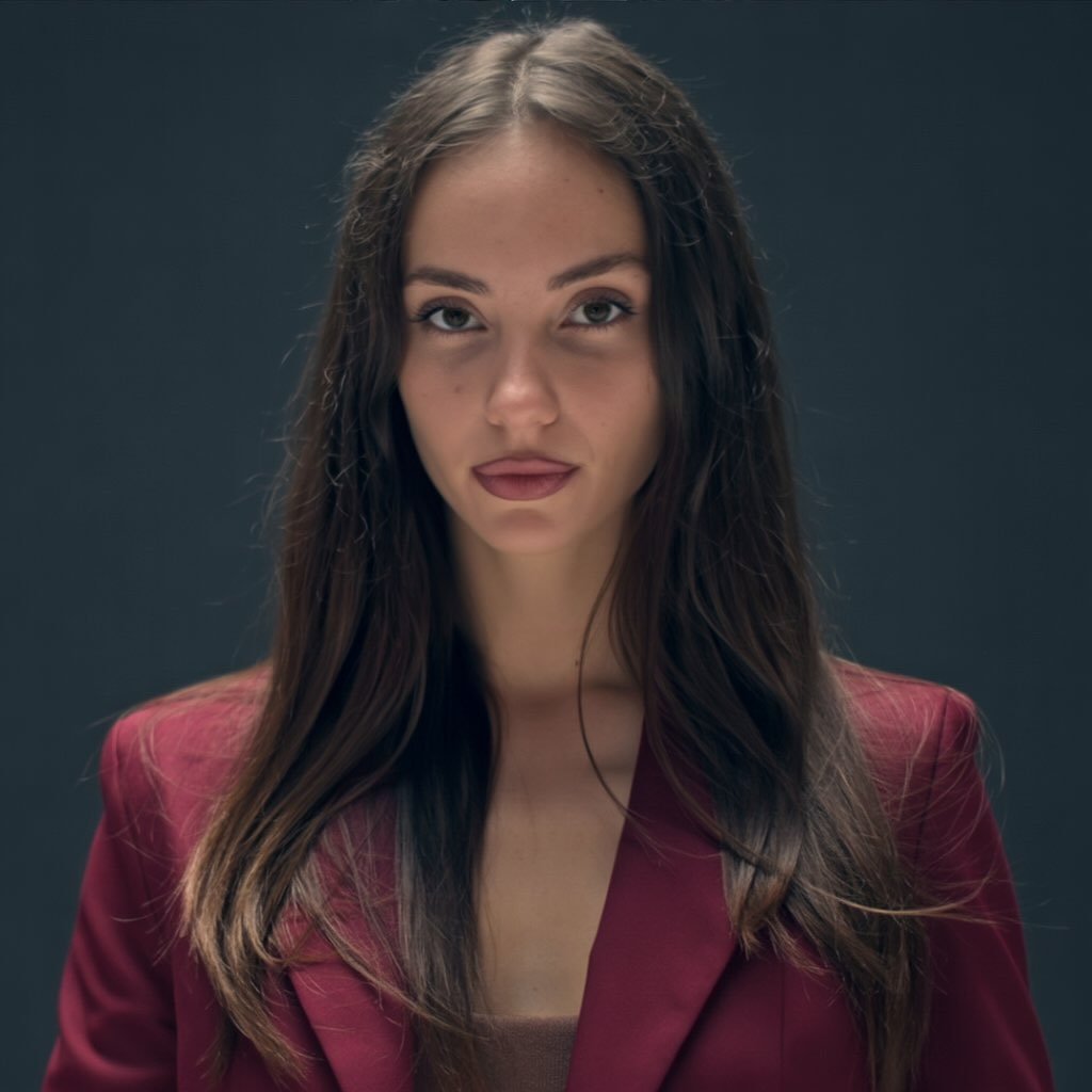 A woman with blonde bob hair in a grey coat and beige turtleneck stands against a neutral backdrop. She has a serious expression, conveying elegance.