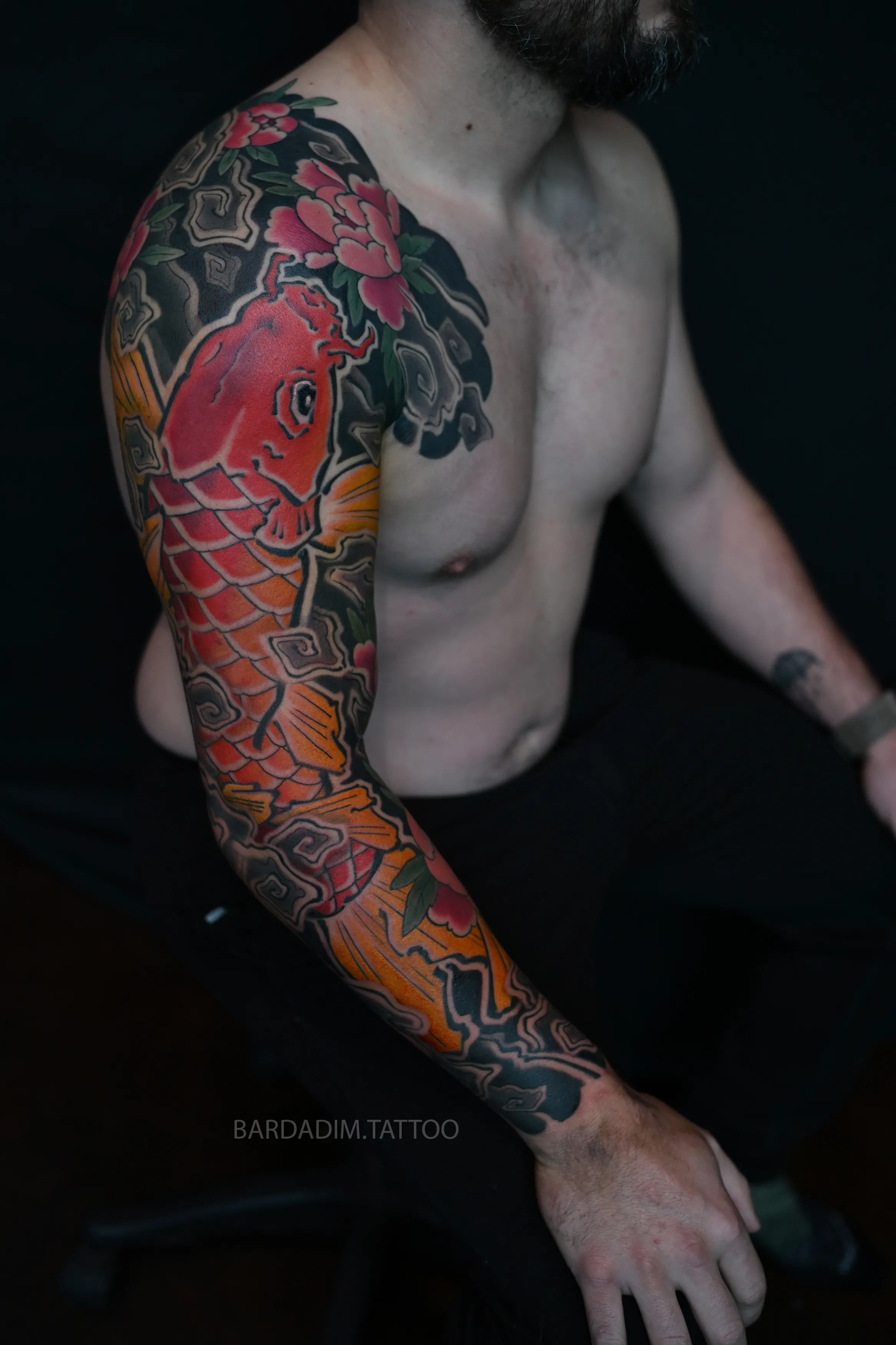 Three-quarter view with the arm bent: the orange koi holds its line from the shoulder to the forearm across the elbow, with a pink peony and the botan-giri border above it.