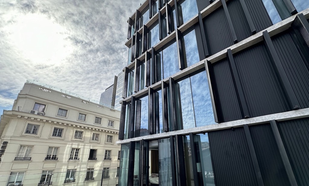 Modern building with glass facade reflecting the sky, contrasting with an older architectural structure nearby.