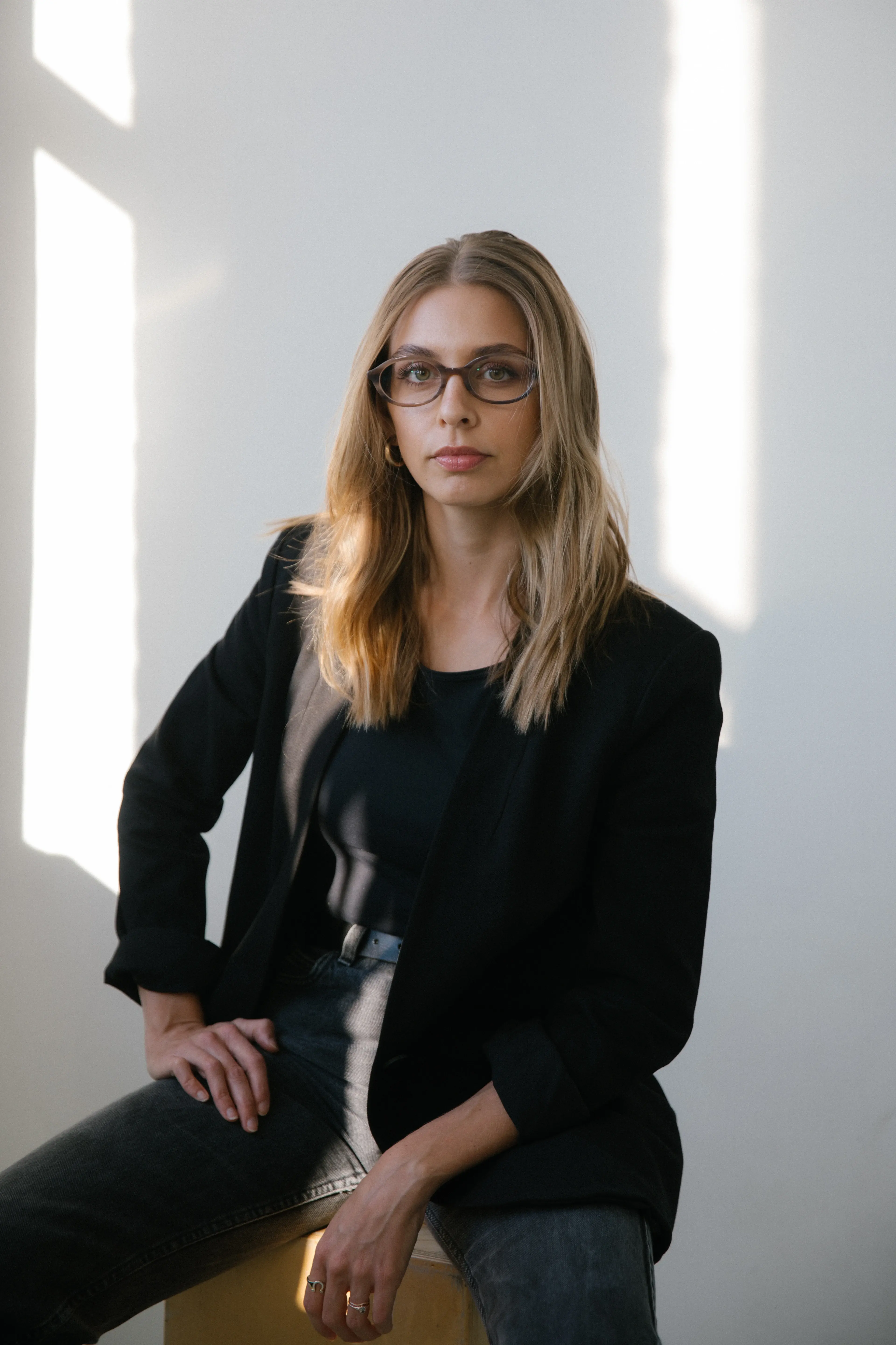Sawyer, a person with long, light brown hair and glasses sits confidently on a stool in front of a white wall, wearing a black blazer and jeans, with sunlight casting shadows on the surface.