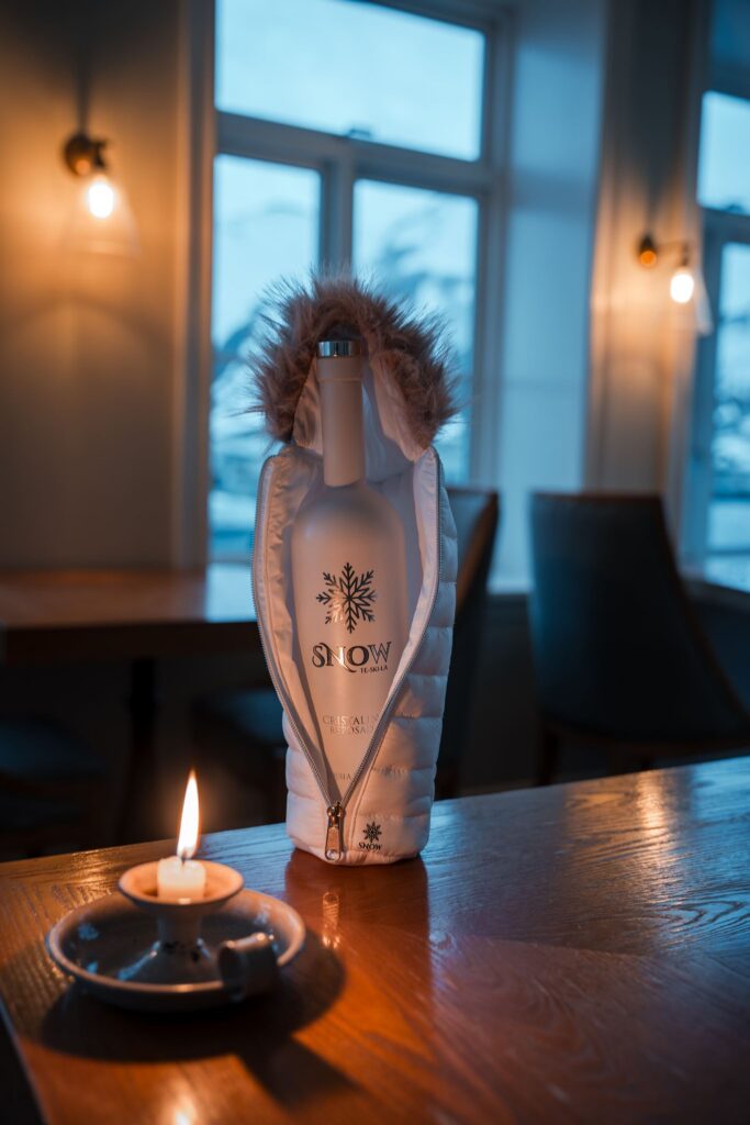 Snow Tequila bottle in a miniature white hooded jacket beside a lit candle on a wooden bar
