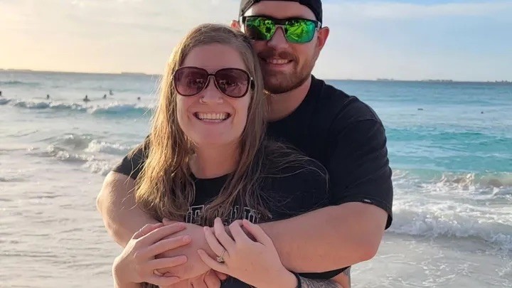 A smiling couple stands on a beach, with the ocean and a sunset in the background. They are embracing happily.