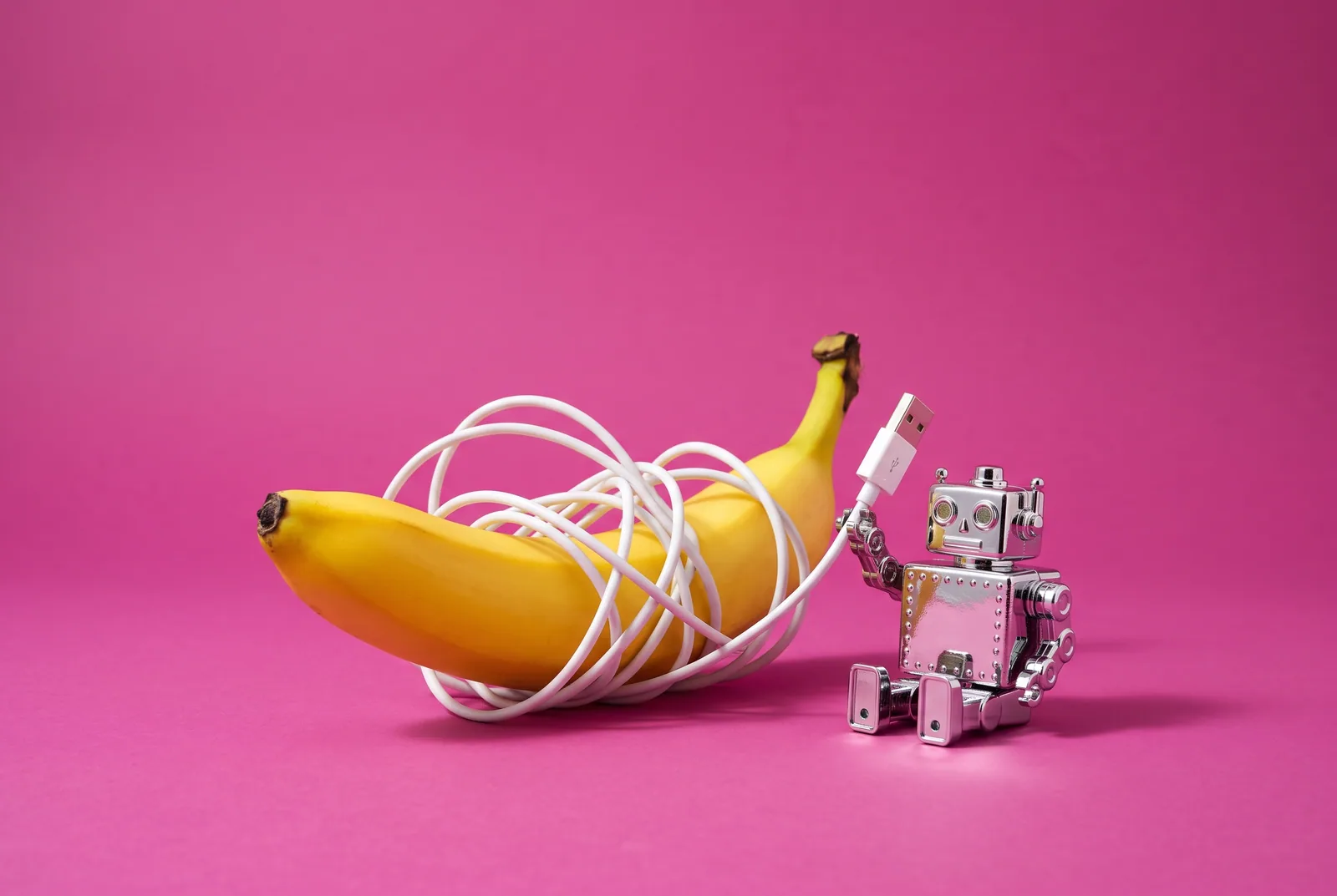 A banana tangled in a charging cable held by a chrome toy robot, signs of AI porn addiction