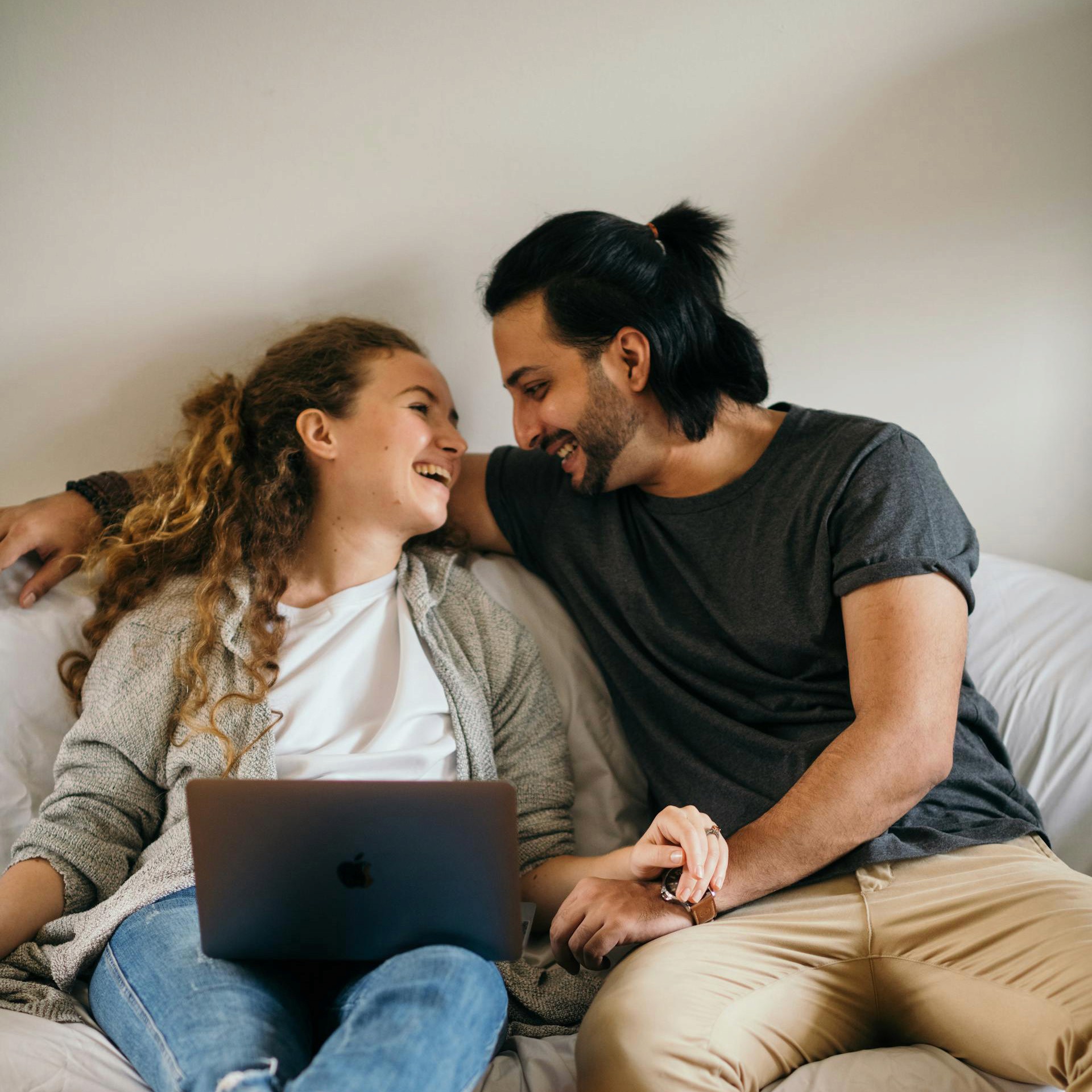 A couple is sitting together, leaning against a headboard. The woman, with curly hair, is holding a laptop in her lap while smiling and looking up at the man, who has long dark hair pulled back in a ponytail and is also laughing. They are holding hands.