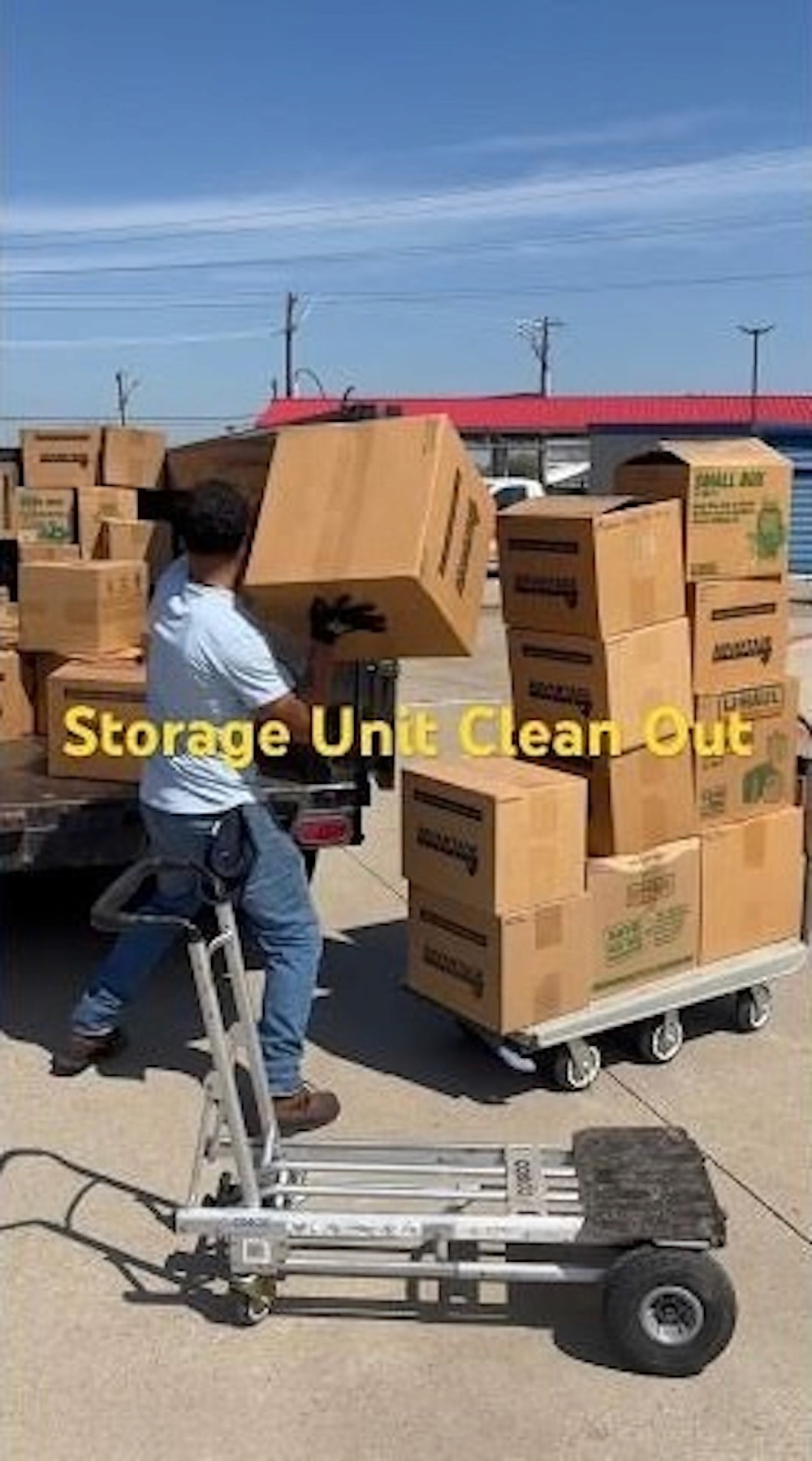 Man loading cardboard boxes onto a cart in a sunlit storage facility. The words "Storage Unit Clean Out" overlay the image, conveying a sense of organization and activity.