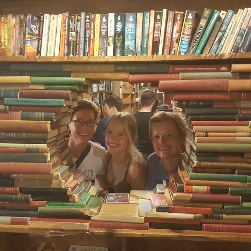 Three people smile through a hole made of stacked books, with a background of filled bookshelves and more people browsing.
