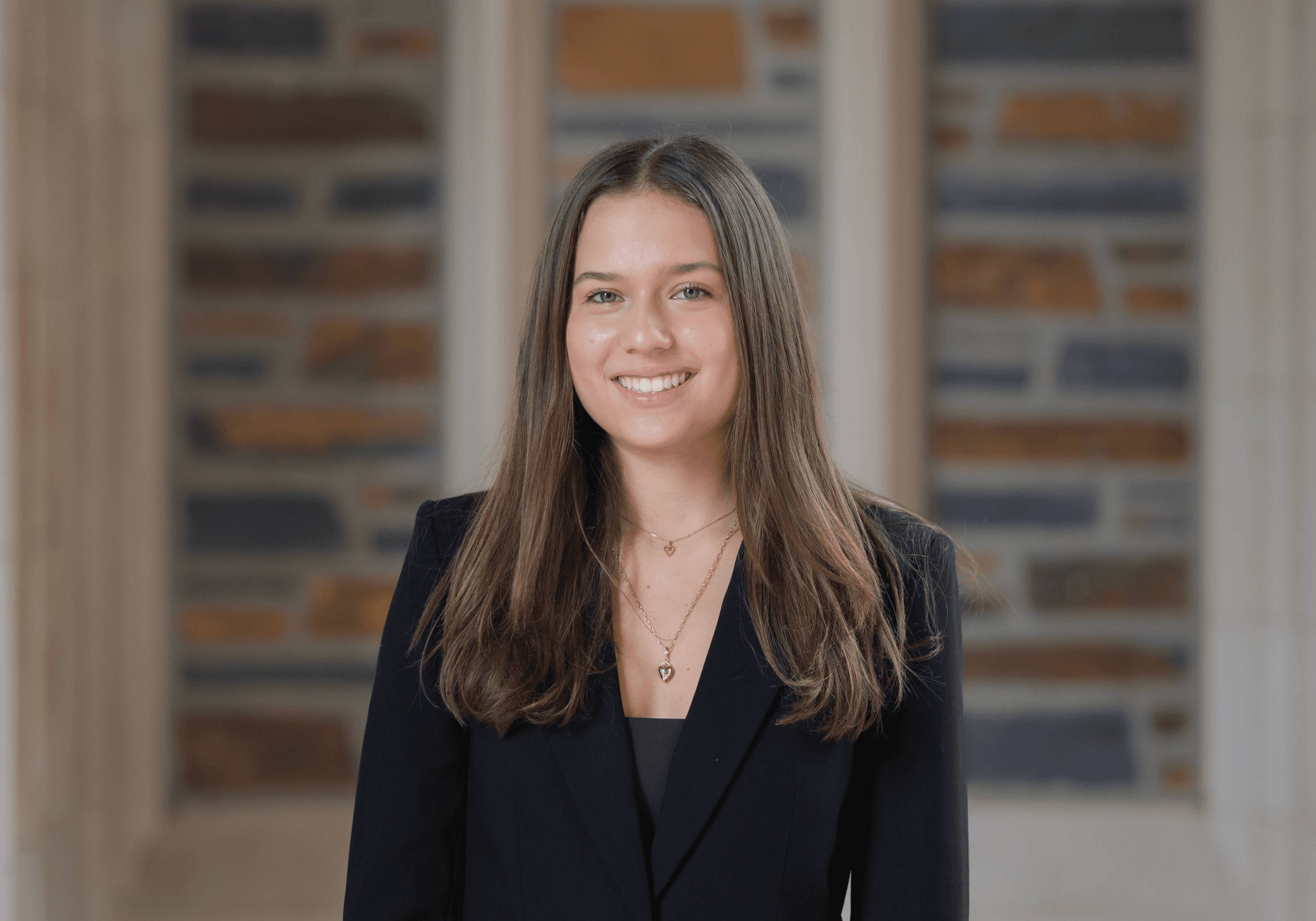 A woman with long brown hair is wearing a black blazer and standing in front of a background featuring a geometric pattern of gray, brown, and blue stone bricks, illuminated by soft natural light.
